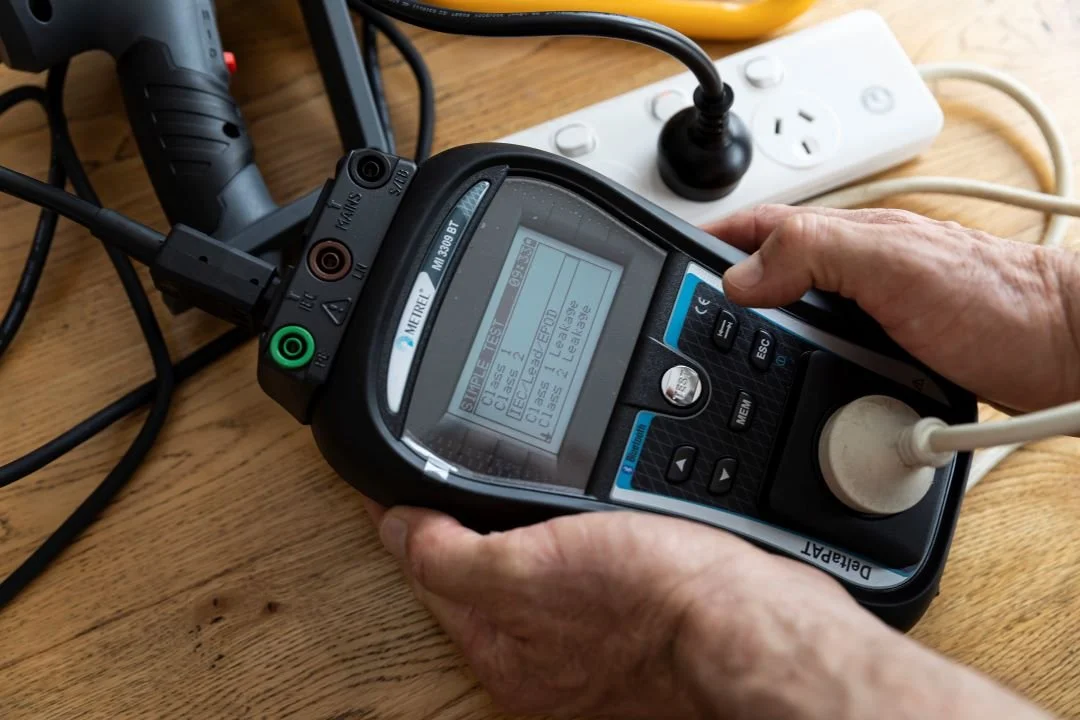 A person holding a device with a digital screen connected to multiple cords and plugged into a power strip on a wooden surface.