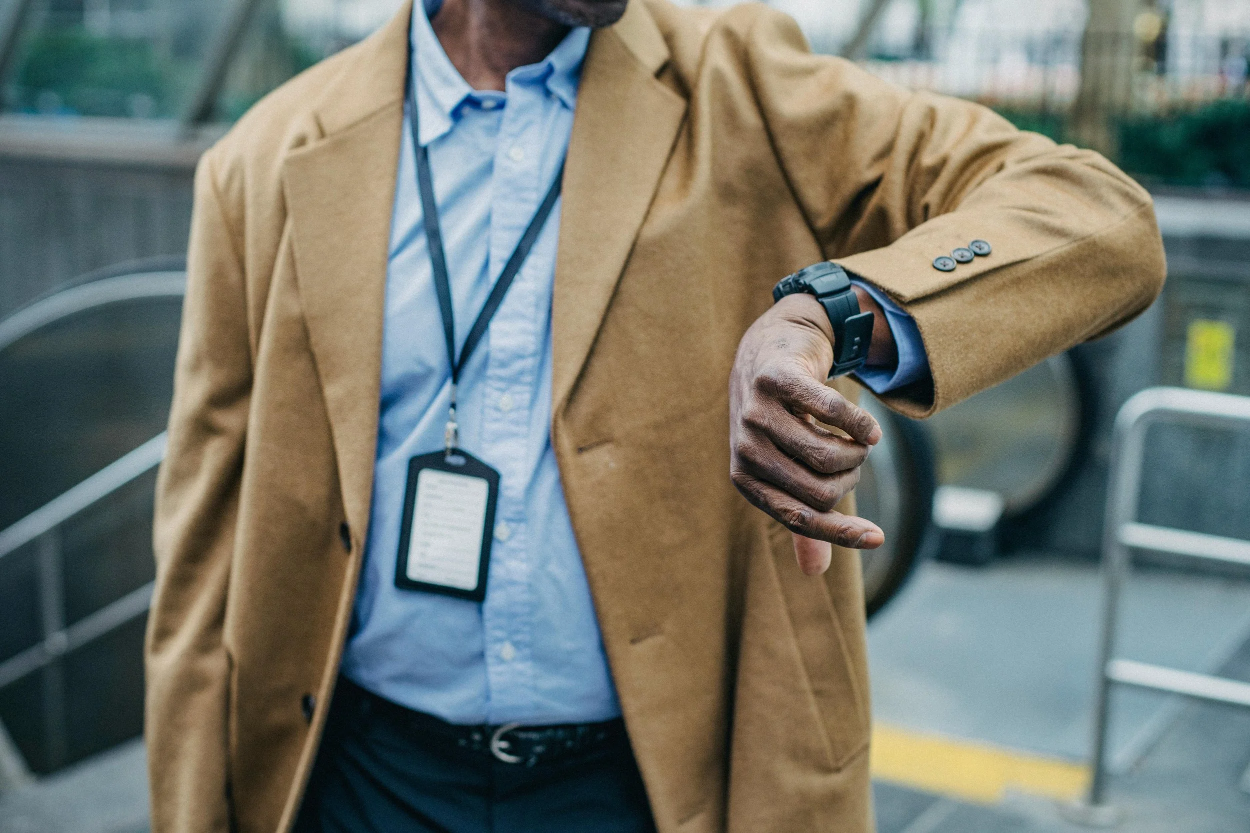 A man wearing a tan blazer, light blue shirt, and lanyard with an ID badge, looking at his wristwatch outdoors near an escalator.