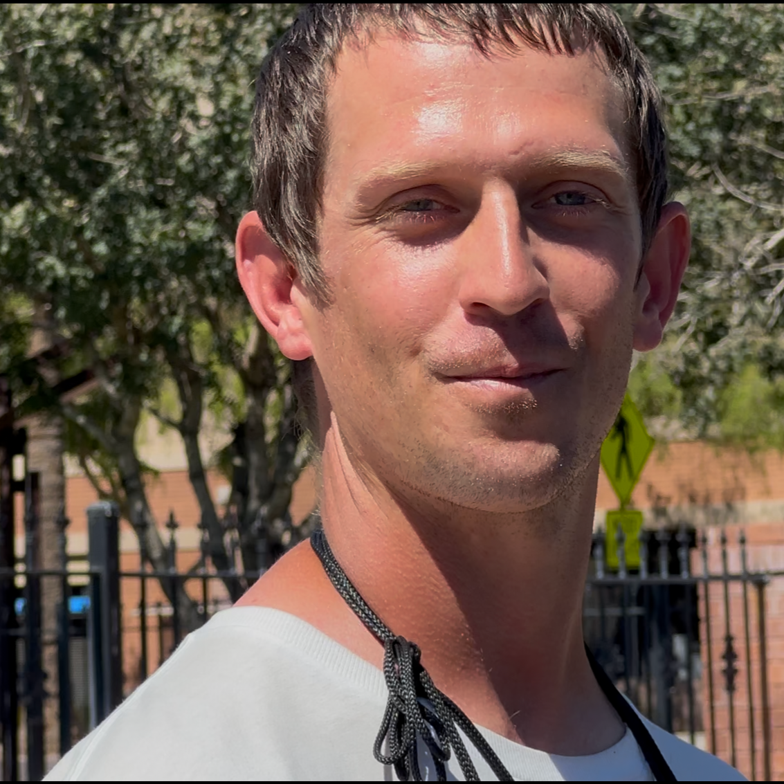 A young man with short brown hair and light skin outdoors, smiling softly, wearing a white shirt and a black lanyard, with trees and a fence in the background.