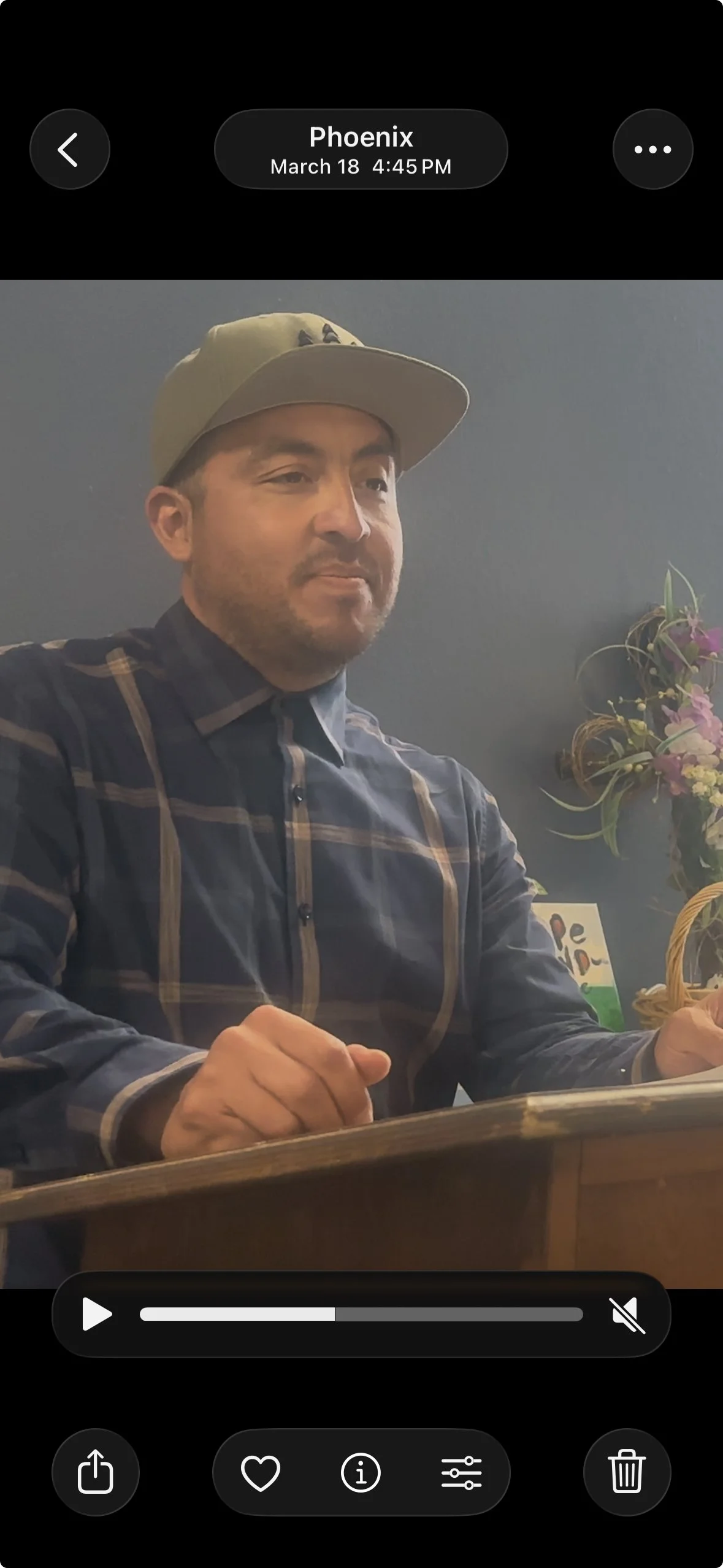 A man is sitting at a table, wearing a plaid shirt and a baseball cap, with a serious expression. There are flowers and a basket in the background.