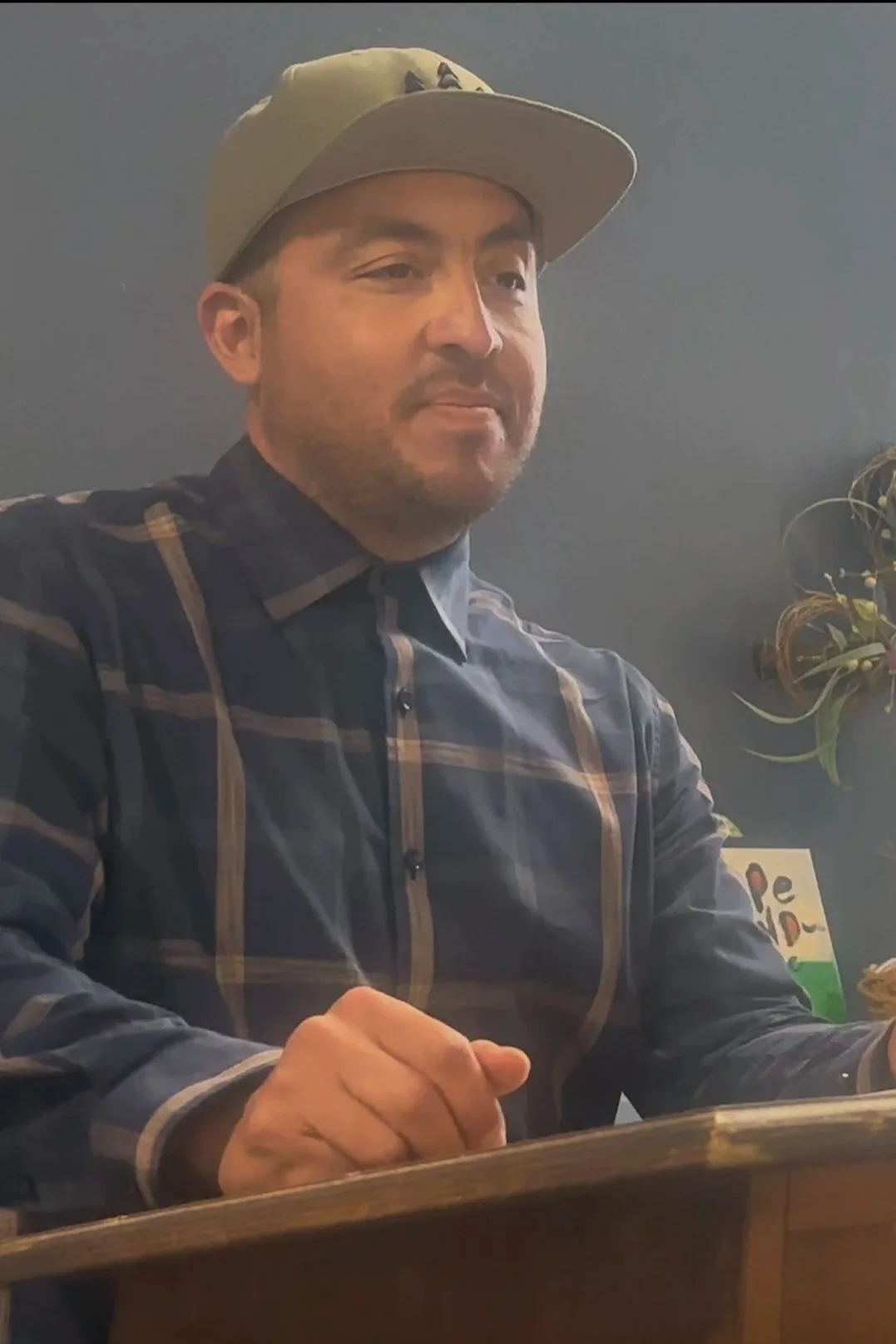 A man with a beard wearing a beige baseball cap and a plaid shirt, sitting at a table.
