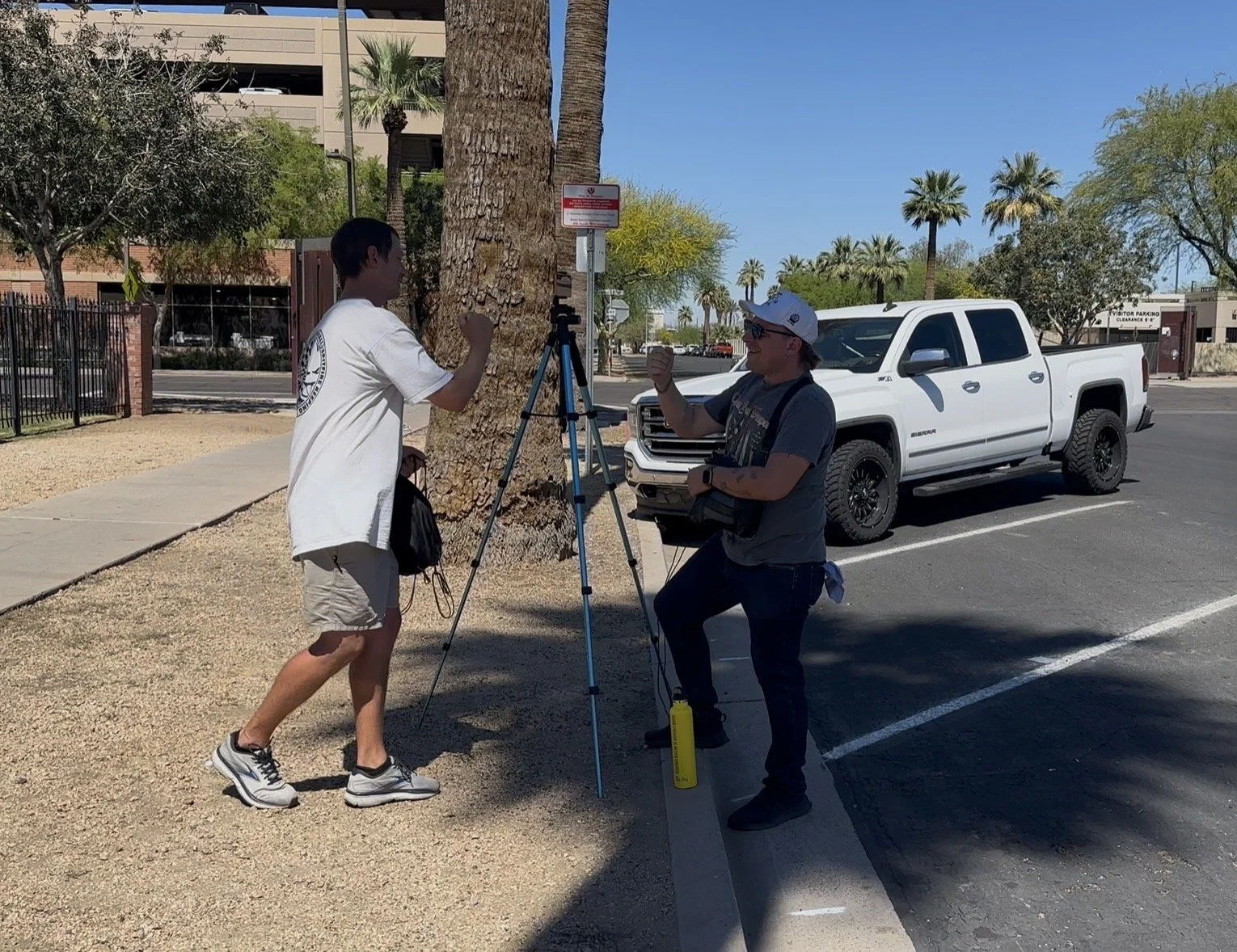 Two people engaging in an activity with a tripod and camera equipment beside a large tree in a parking lot. One person is playing a punching game while the other raises a fist in a mock boxing stance.