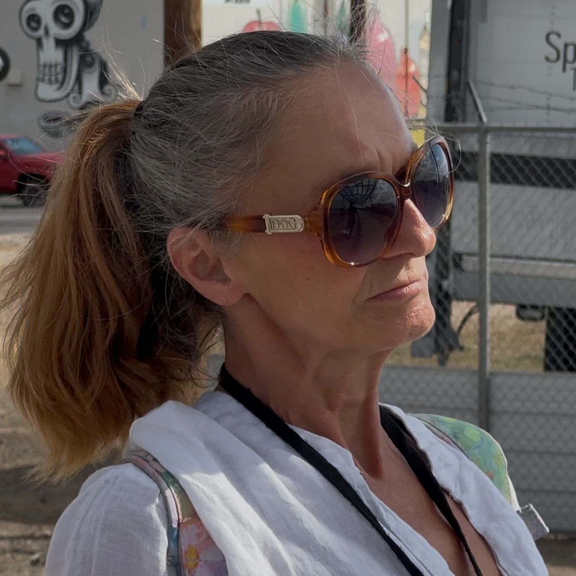 A woman with light skin and reddish-brown hair tied back in a ponytail, wearing large round tortoiseshell sunglasses, a white shirt, and a black lanyard, outdoors near a chain-link fence and a trailer.