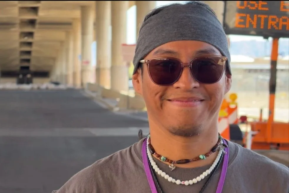 Smiling man wearing dark sunglasses and a gray headband outdoors near a road with construction signs.