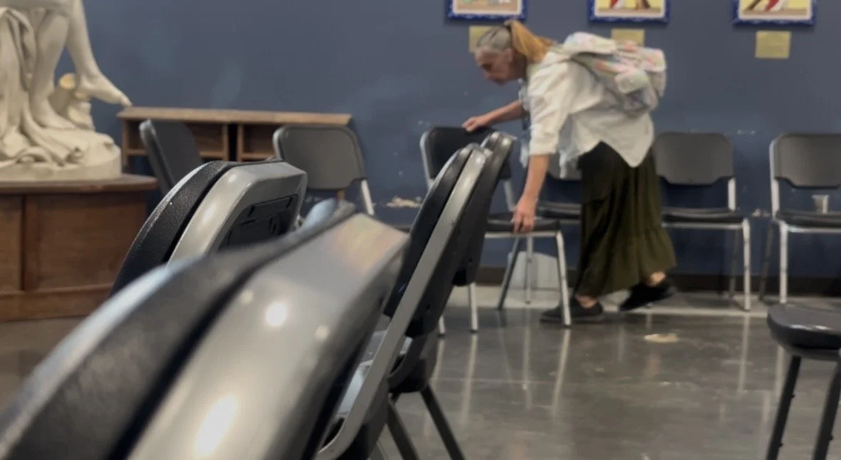 A woman wearing a light-colored shirt and long skirt arranging chairs in a room with blue walls and artwork on the wall.