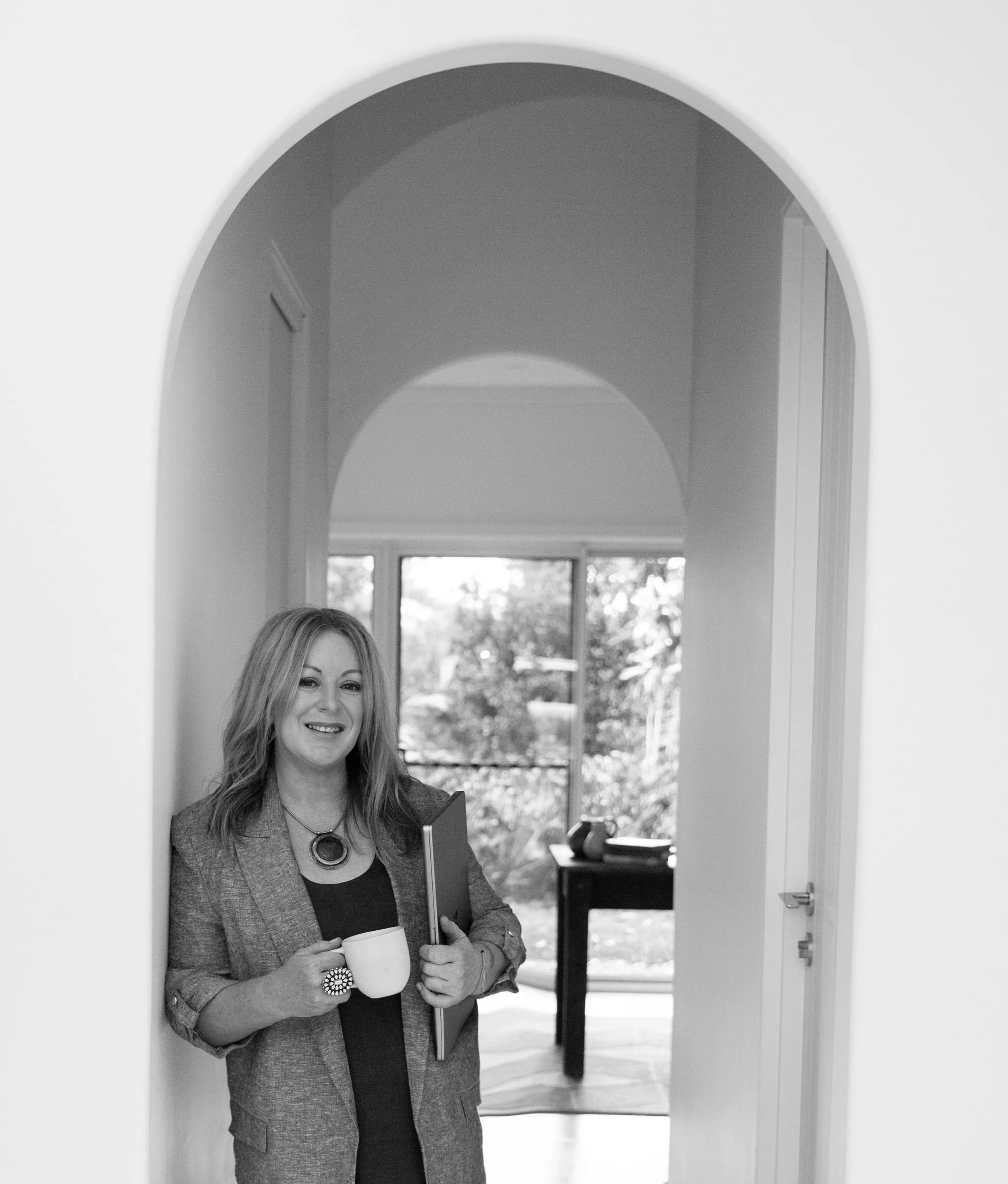 A woman with long hair smiling while holding a coffee mug and a tablet, standing in a bright room with a large arched window and outdoor trees visible in the background.