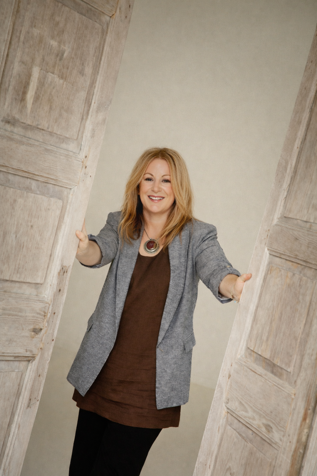 A woman with strawberry blonde hair smiling, wearing a grey blazer and brown dress, standing in a doorway with rustic wooden doors.