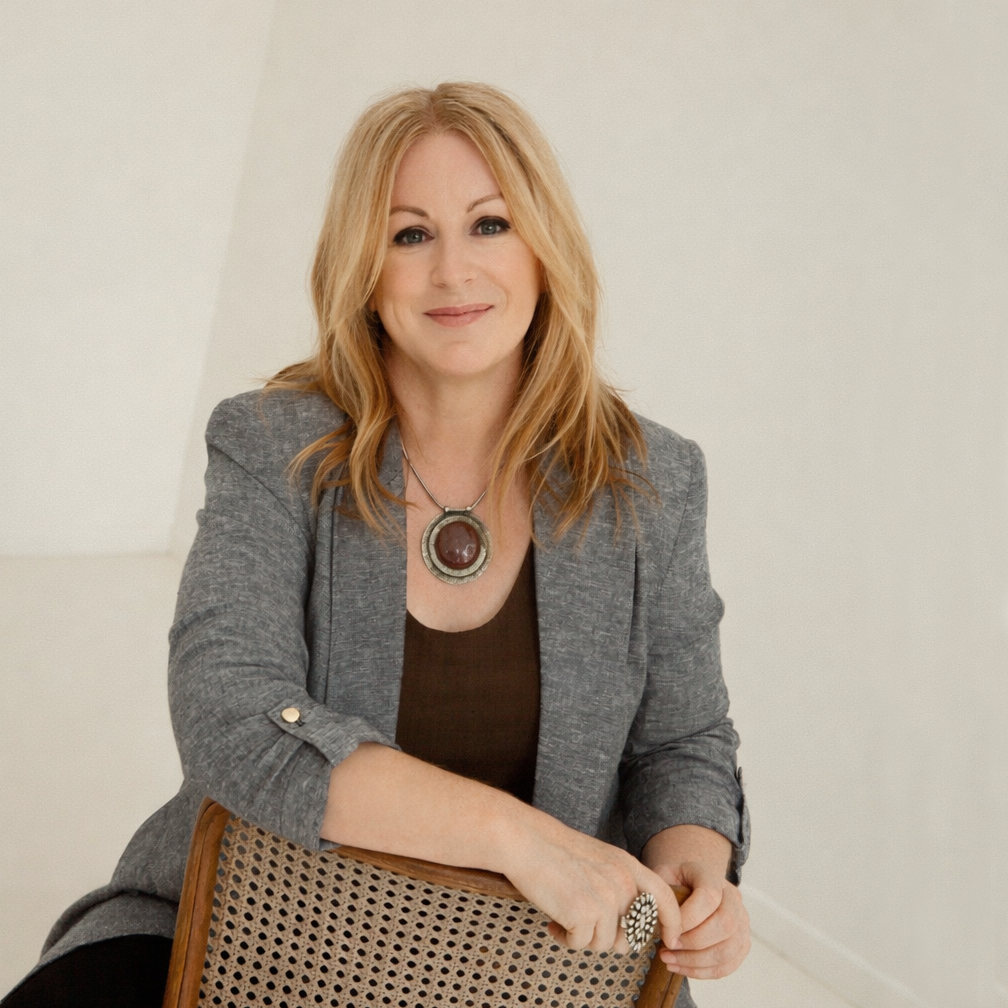 A woman with strawberry blonde hair wearing a grey blazer, black top, and a large necklace, sitting on a chair with a woven back. She is smiling and facing the camera against a plain white wall.
