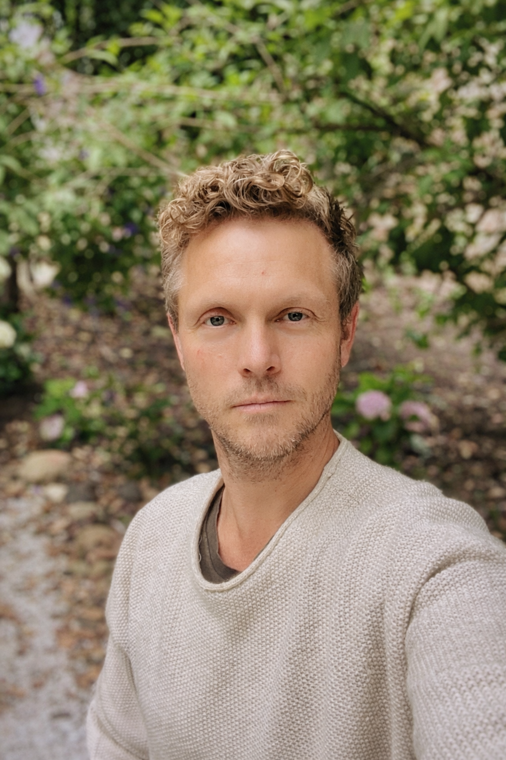 A man with curly blonde hair and blue eyes taking a selfie outdoors. He is wearing a light-colored textured sweater and is standing in front of a background with green foliage and blooming plants.