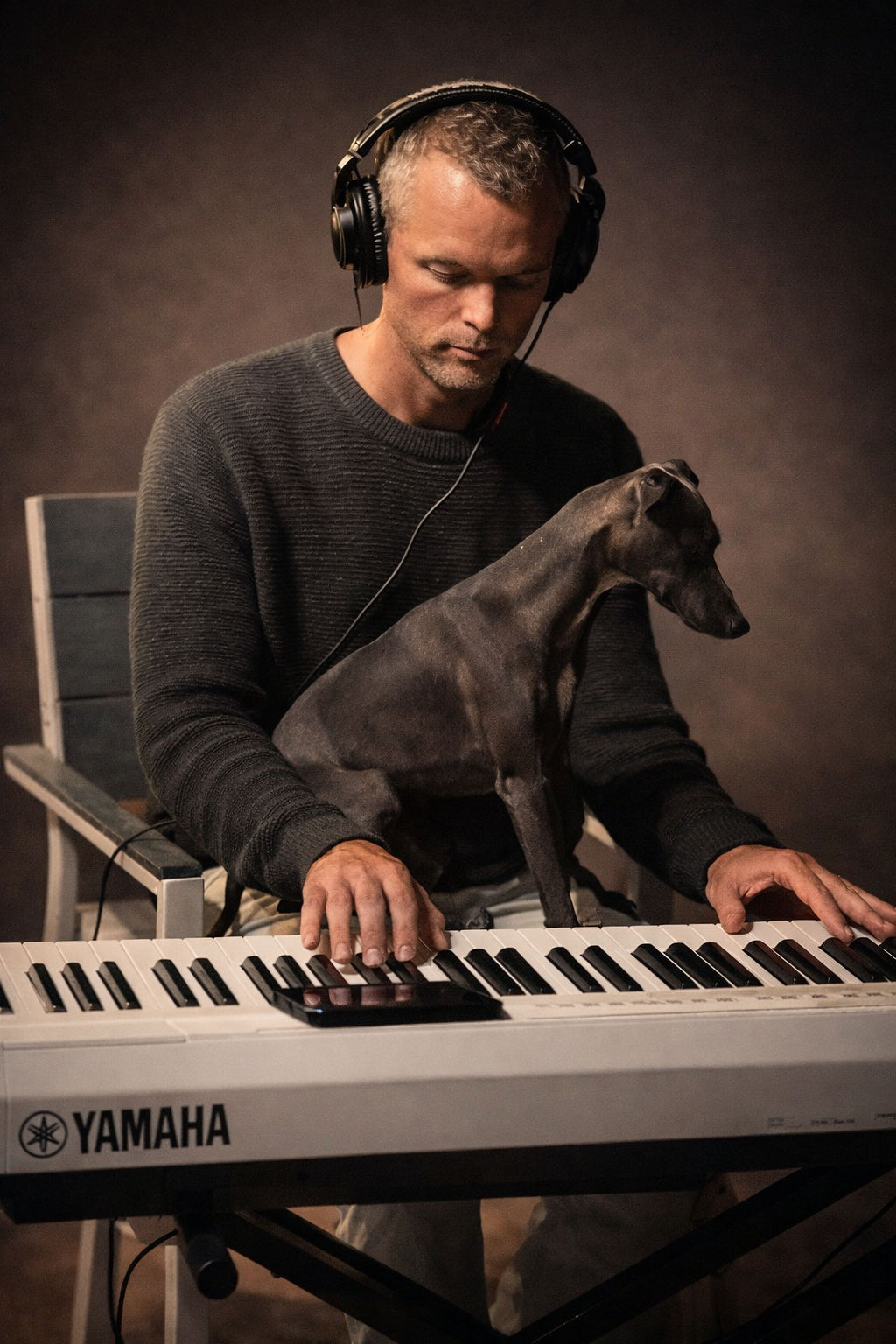 A man with gray curly hair, wearing a dark sweater and headphones, playing a Yamaha keyboard with a black dog sitting on his lap, during a music session in a dimly lit room.