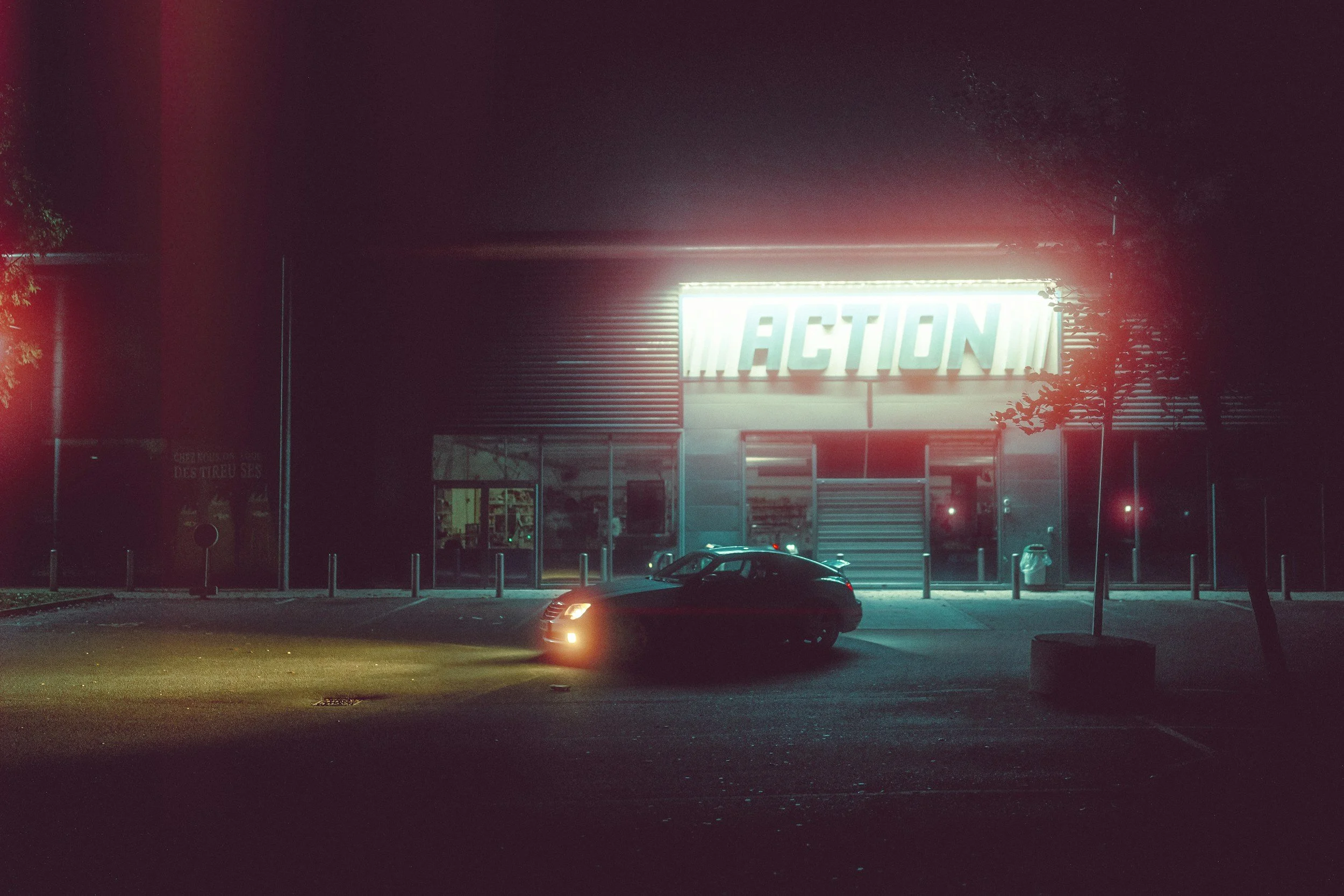 Nighttime scene of a parking lot with a single black car, illuminated by the car's headlight, in front of a building with a bright neon sign that says 'ACTION'.