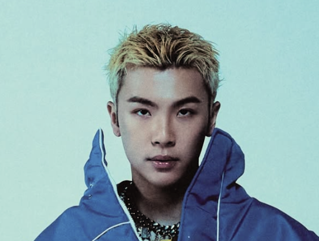 Jeune homme avec des cheveux blonds et une veste bleue, regardant droit vers la caméra.