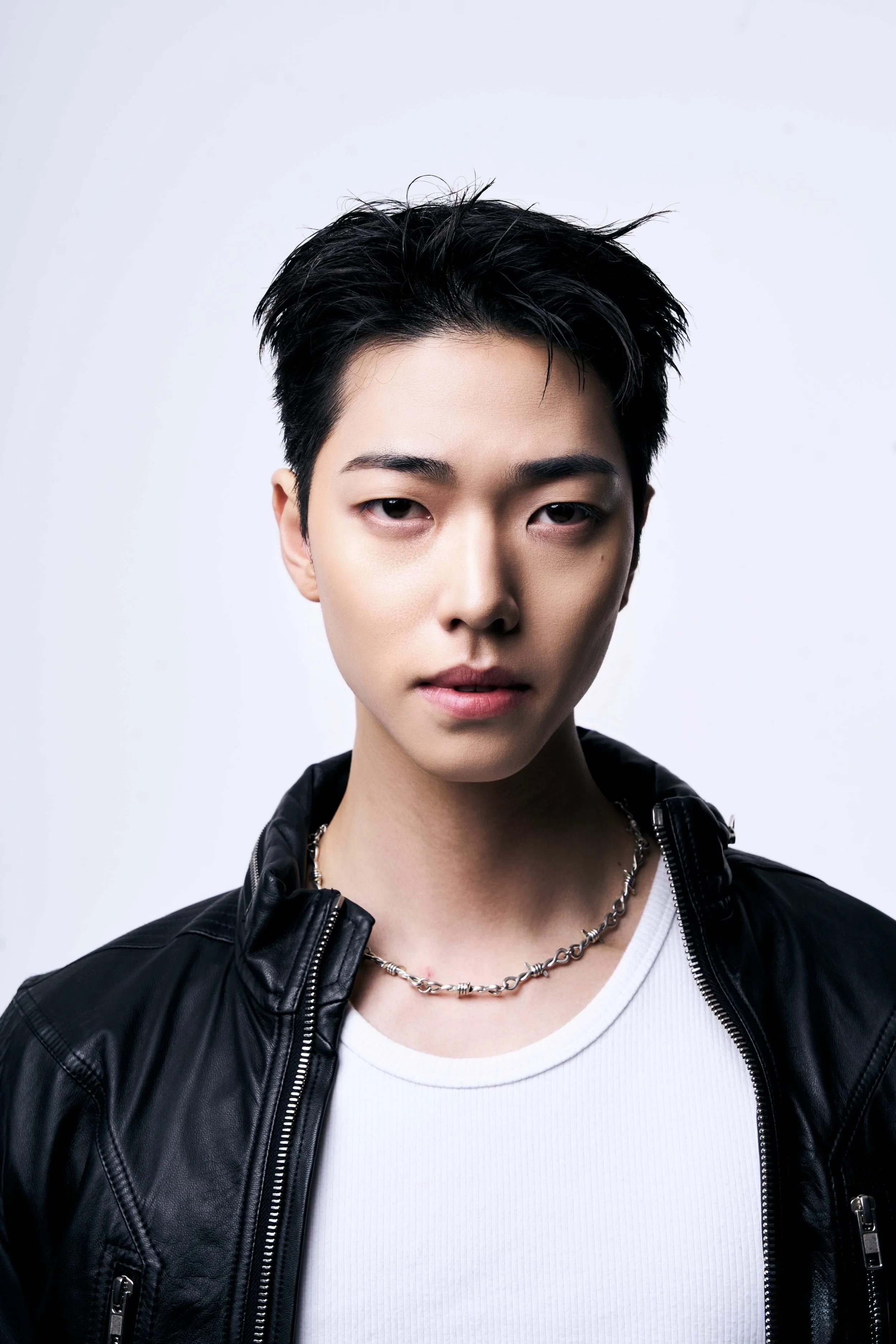 Un jeune homme avec des cheveux noirs, portant une veste en cuir noir et une chaîne, regardant l'objectif, sur fond blanc.