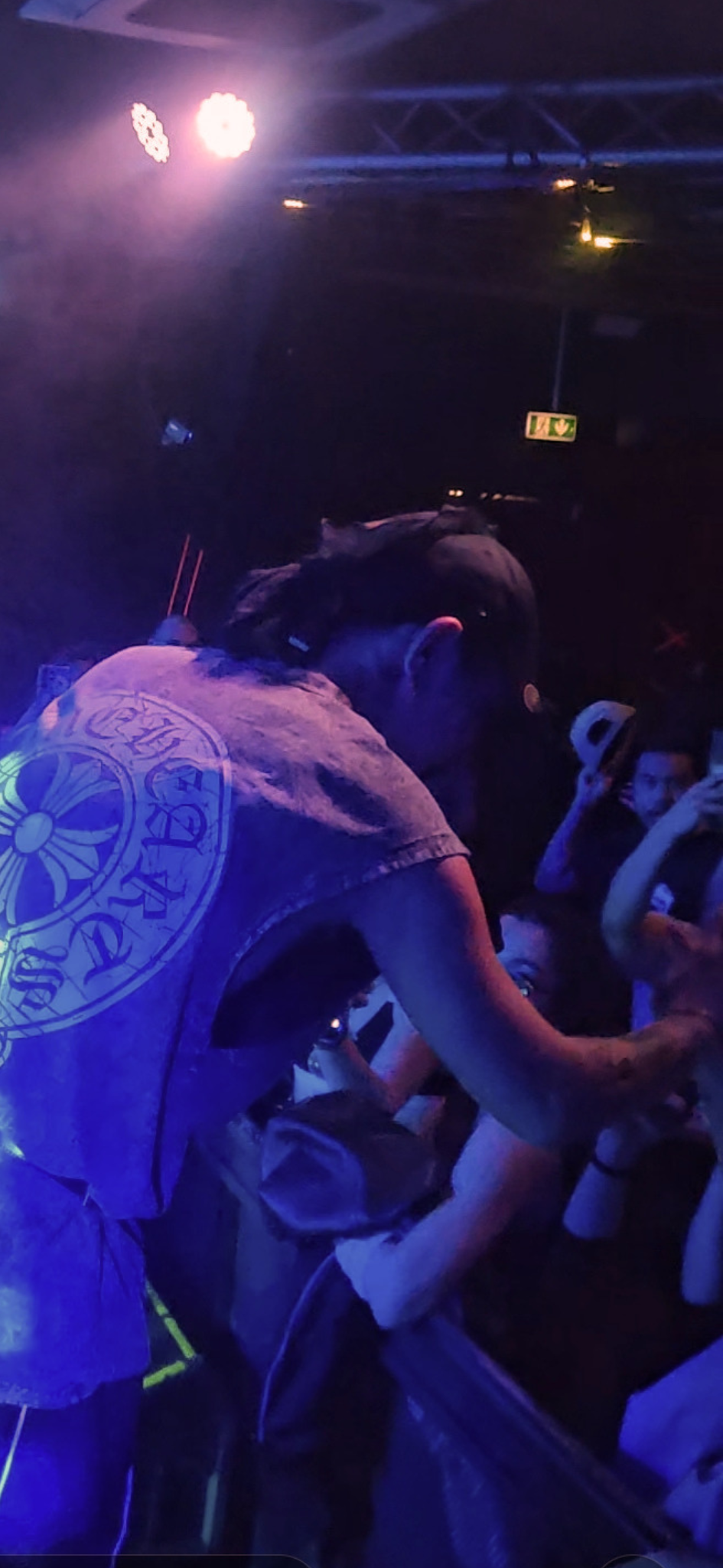 Une personne en t-shirt avec un logo flottant sur le dos rejoint un groupe de jeunes fans lors d'un concert ou d'un événement, avec une ambiance sombre et des lumières colorées.