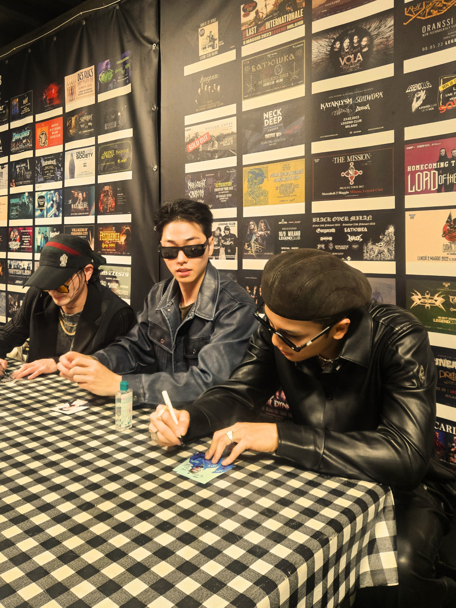 Trois personnes assises à une table à carreaux jouant à un jeu, avec un mur en arrière-plan couvert de posters d'événements musicaux