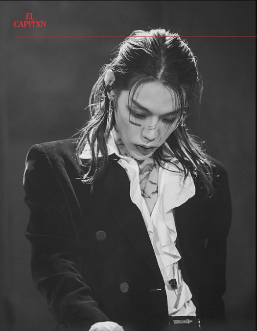 Une femme avec des cheveux brun foncé, portant une veste noire, une chemise blanche froissée, et un micro attaché à sa chemise, apparaissant perdue ou concentrée. Textes rouges en haut à gauche : "EL CAPITXN".