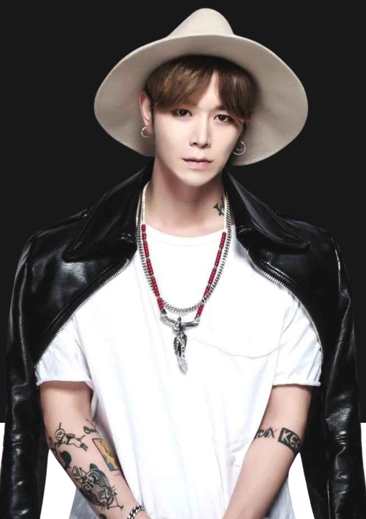 Une personne portant un chapeau beige, un t-shirt blanc, une veste en cuir noir, plusieurs colliers, des tatouages sur les bras et porte des boucles d'oreilles. Fond noir.