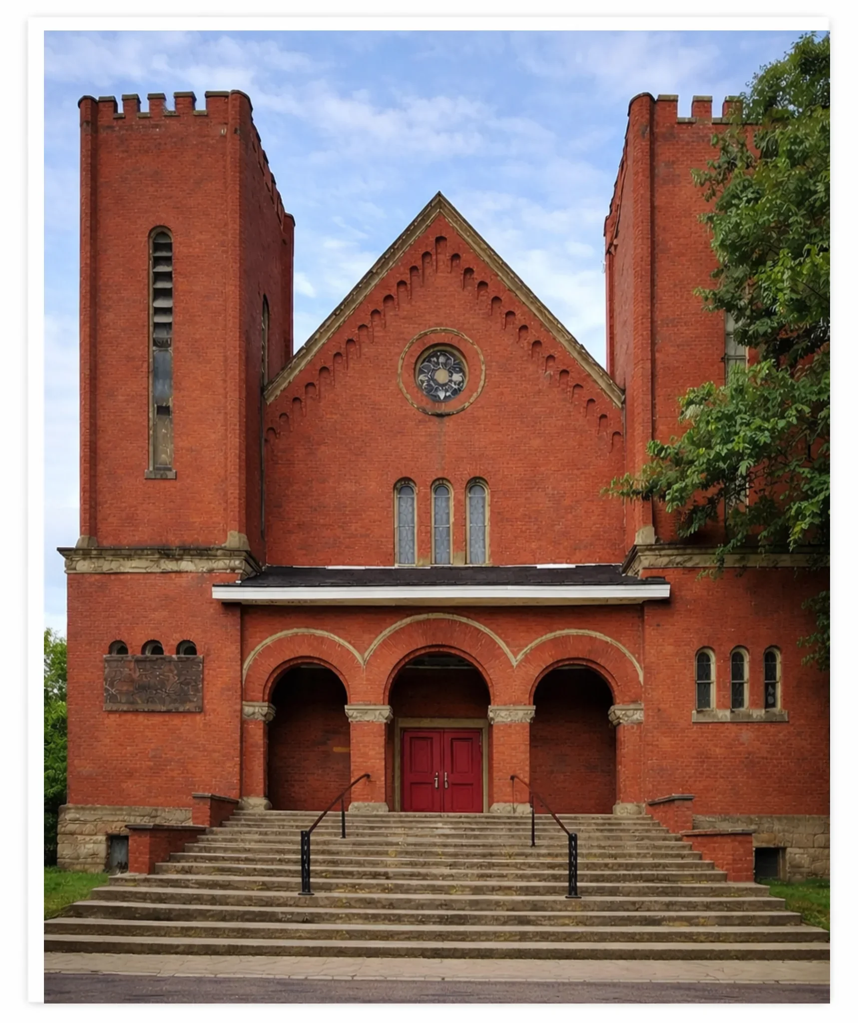 A red brick church with two towers, arched entrances, and stairs leading up to the doors, surrounded by greenery.