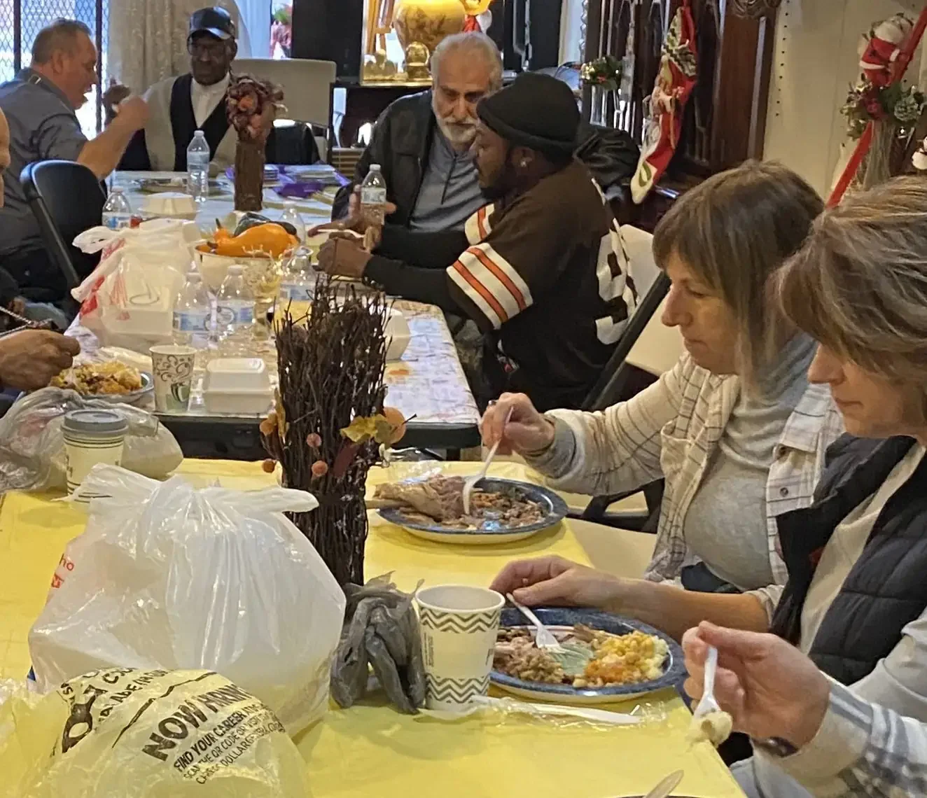 People gathered around a long dining table enjoying a meal, with food, drinks, and plastic bags on the table, in a decorated room with holiday decorations.
