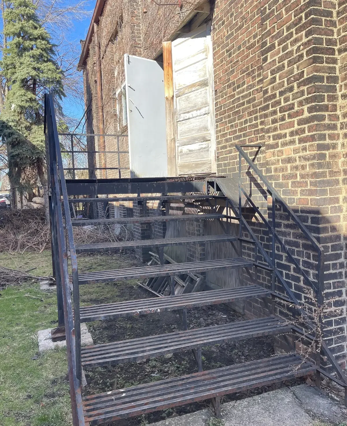 Metal outdoor staircase leading to a door on a brick building with a wooden panel. The area is outdoors with a tree and some grass nearby, and the sky is blue with some clouds.