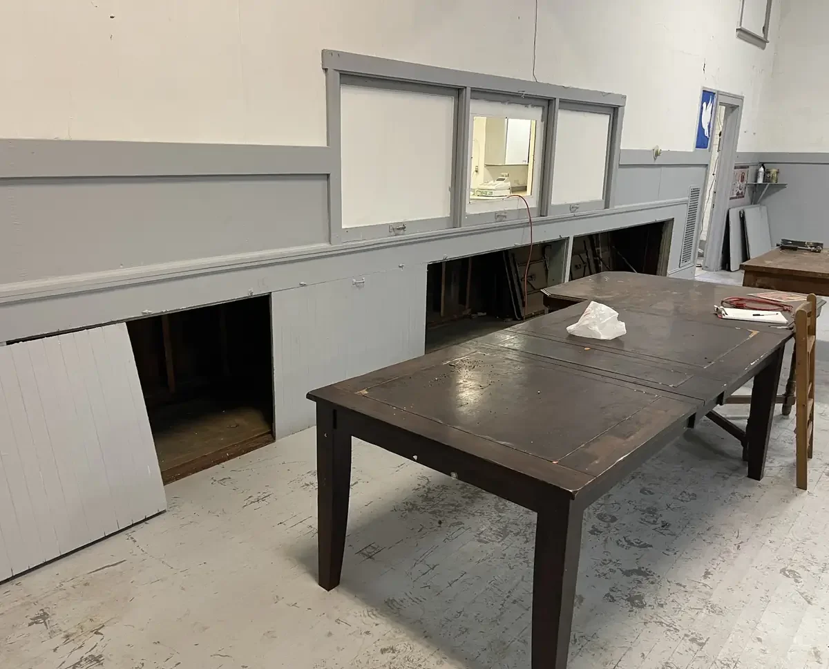 Interior of a room with construction or renovation work underway, featuring a wooden table, open wall panels, and a window with a view into another room.