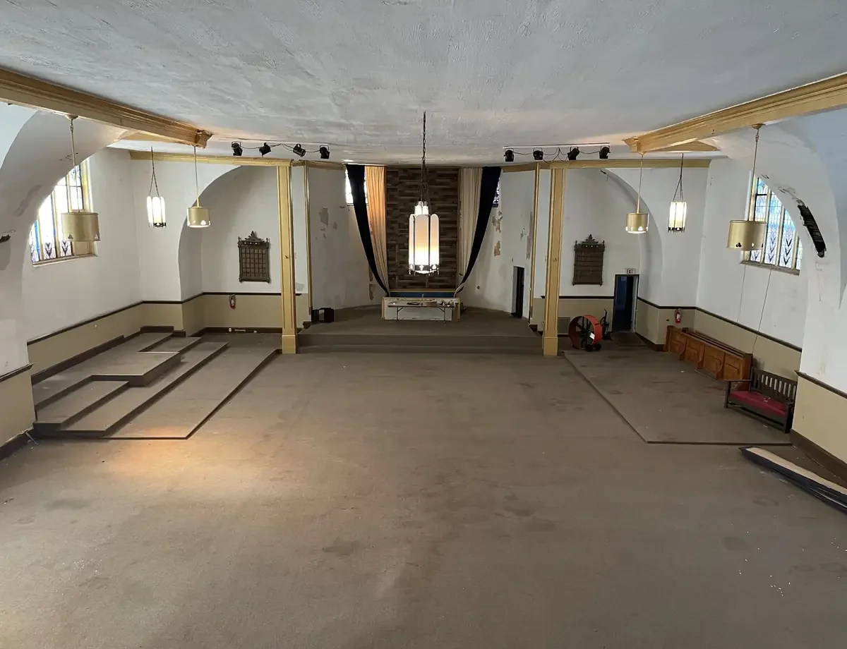 Empty church hall with a small stage at the front, featuring hanging lantern-style lights, stained glass windows, and a few chairs and benches along the sides.