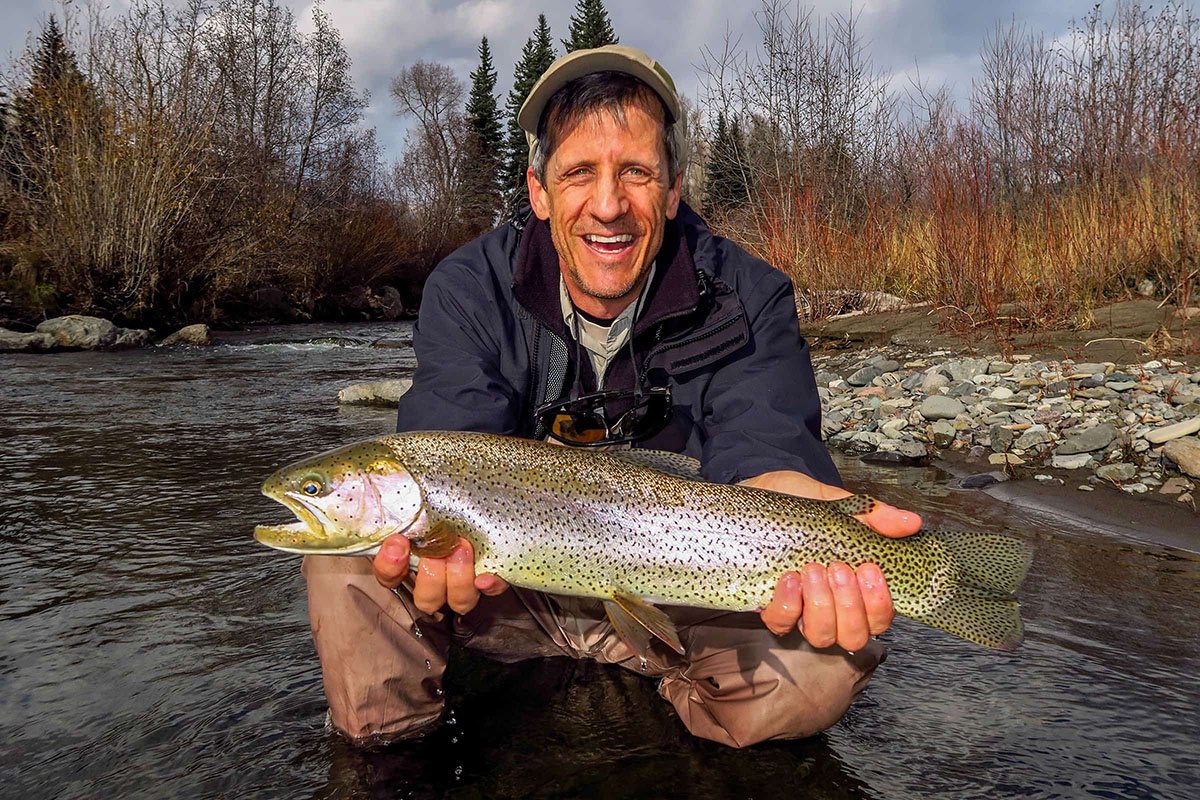 Smiling man kneels in a stream, holding a large fish with both hands; trees and rocks are in the background.