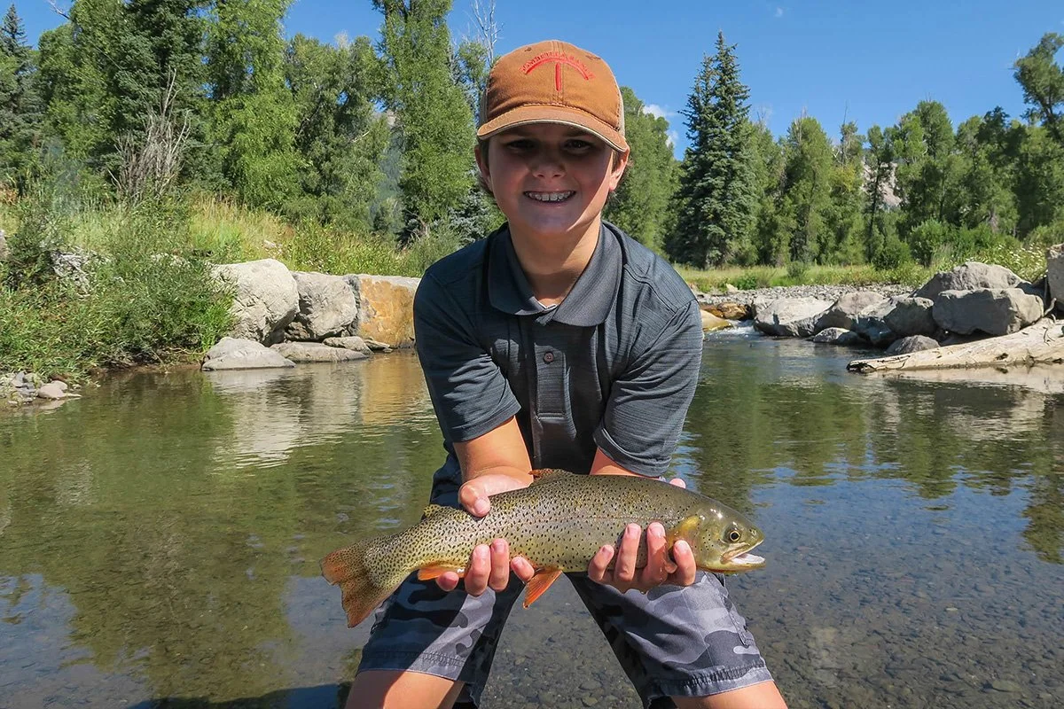 Smiling boy kneels in a shallow stream, holding a large fish with both hands; trees and rocks in the background.