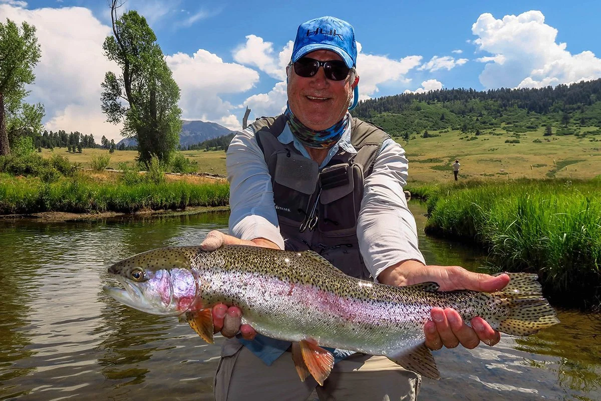 Smiling person in fishing gear holds a large rainbow trout by a clear stream in a grassy, mountainous landscape under a blue sky.