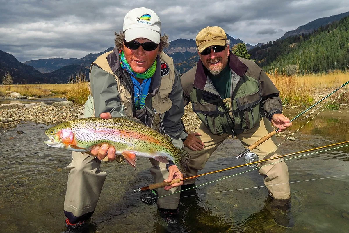 Two fishermen in outdoor gear proudly hold a large rainbow trout in a shallow river with mountains and trees in the background.