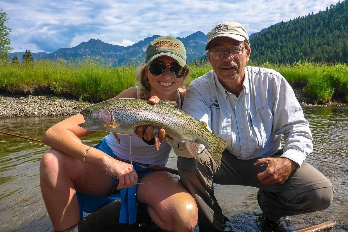 Two people kneel in a stream, smiling, as one holds up a large fish with mountains and trees in the background.