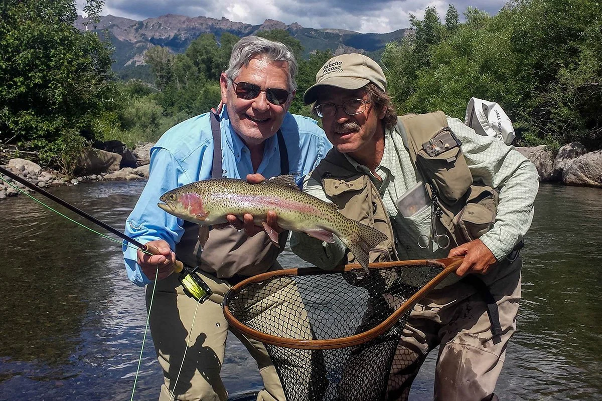 Two men smiling while holding a fishing net with a large trout, standing in a shallow river with trees and mountains in the background.