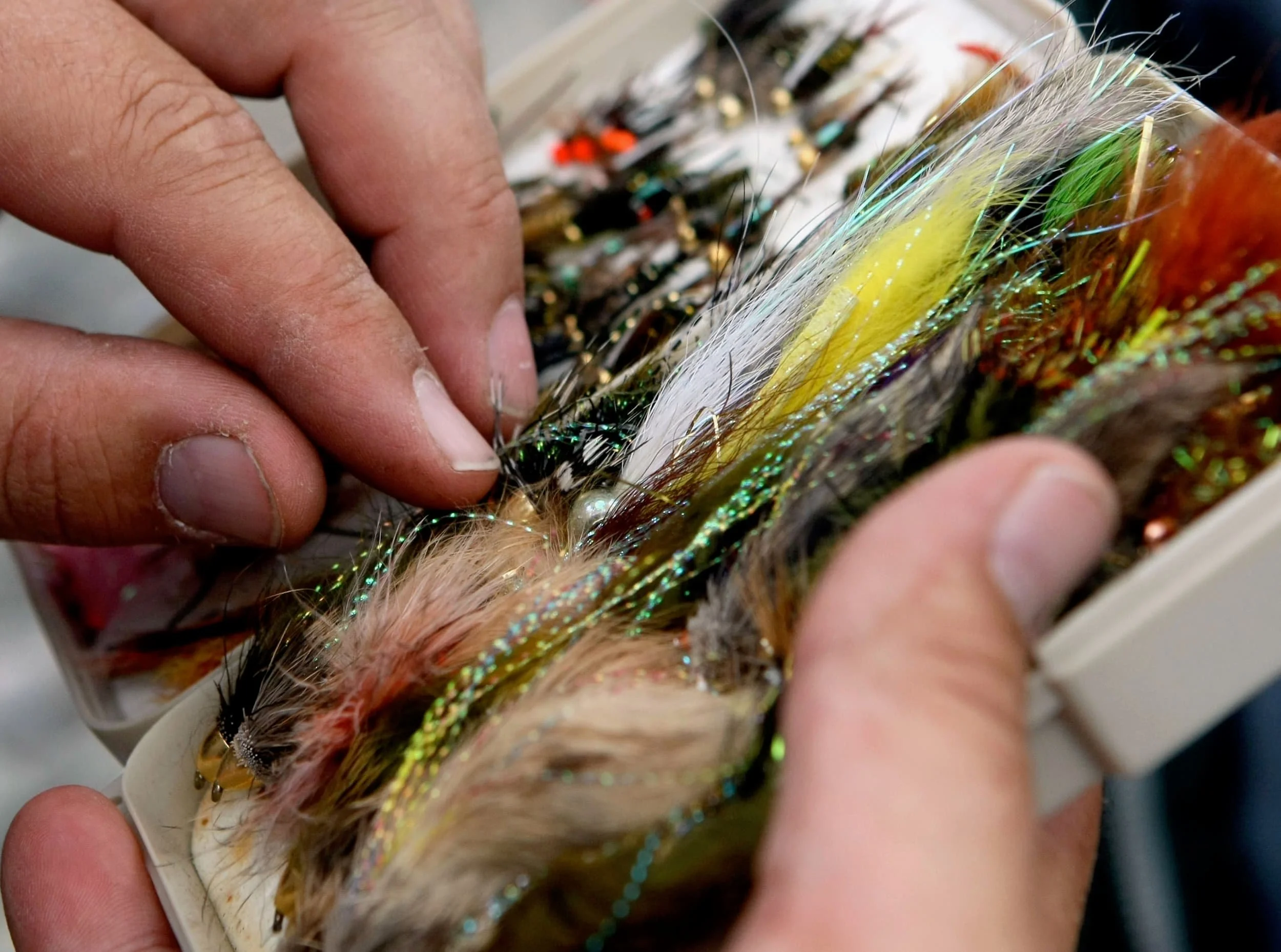 Close-up of hands selecting colorful fishing flies from a small tackle box filled with various artificial lures.