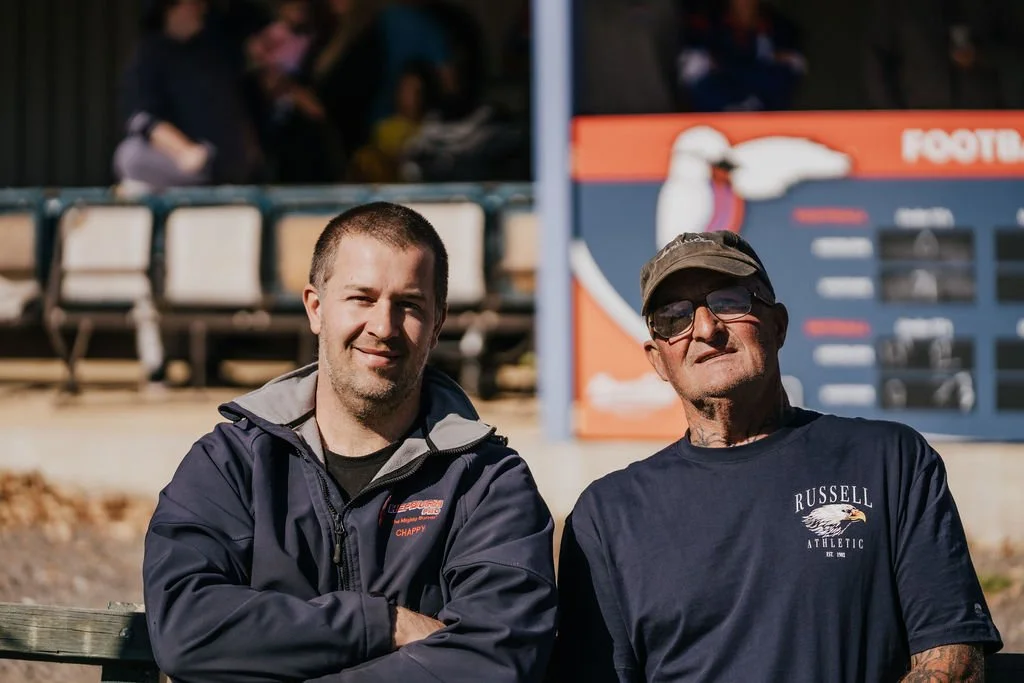 Two Hepburn Football Club Members sitting on a bench in front of the scoreboard