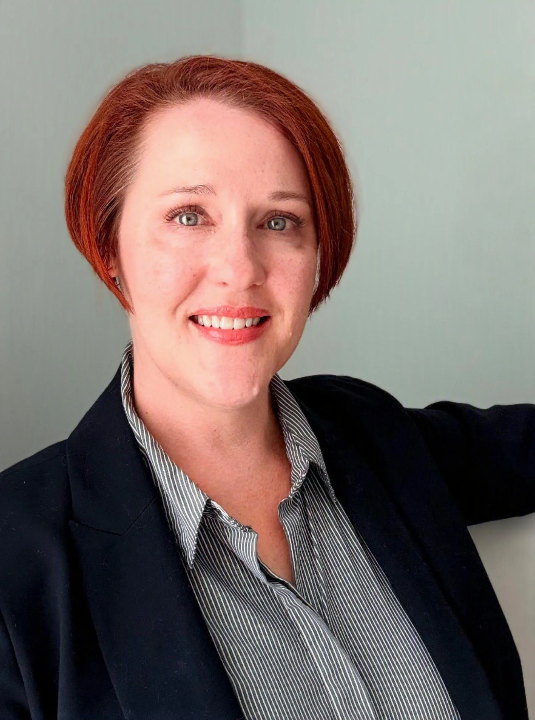 A woman with short red hair, blue eyes, wearing a black blazer and a striped shirt, smiling against a light green background.