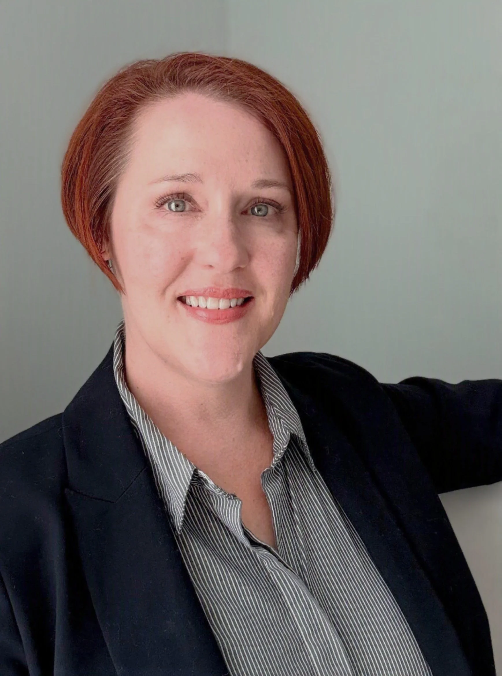 A woman with short red hair, wearing a dark blazer and a striped blouse, smiling.