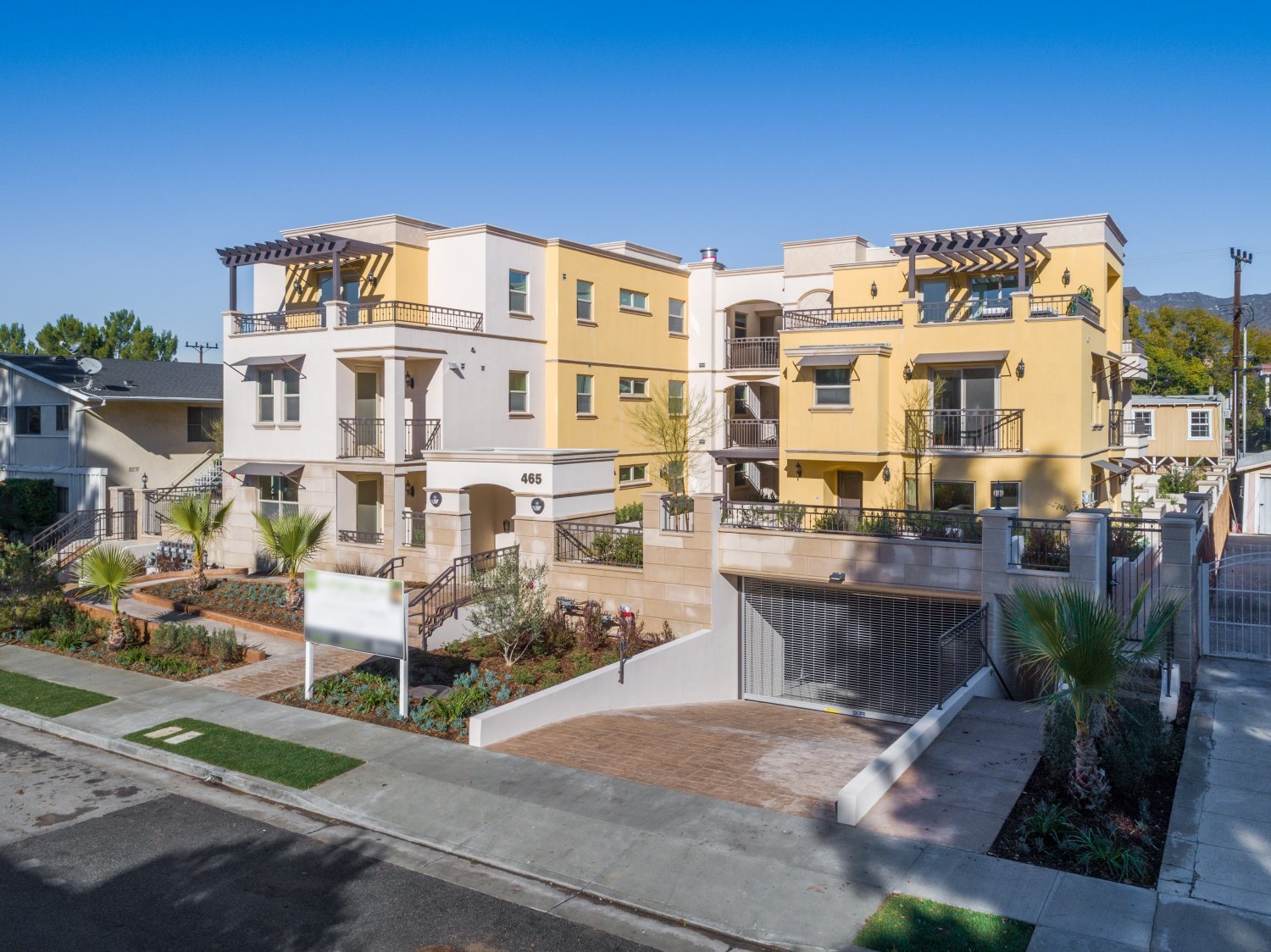 Exterior view of a modern multi-story apartment building with yellow and white facade, balconies, stairs, and landscaped area with palm trees and plants, under a clear blue sky.