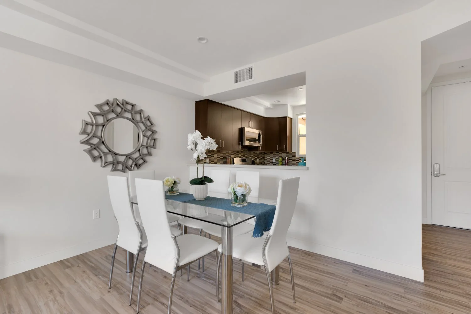 Contemporary dining area with a glass-top table, six white chairs, and floral centerpieces, adjacent to an open kitchen with dark cabinets and mosaic tile backsplash.