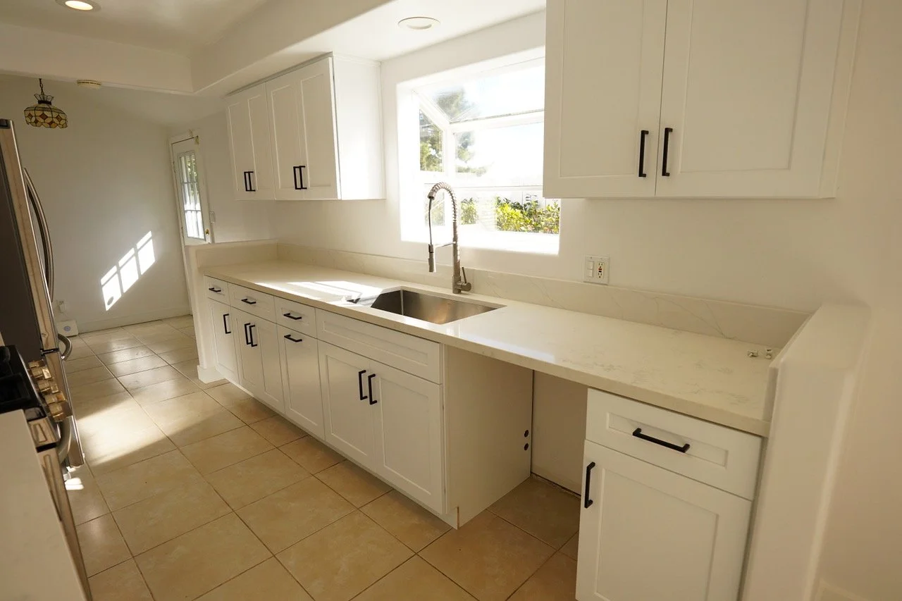 Kitchen with white cabinets, marble countertops, a window above the sink, beige tiled floor, and sunlight streaming in.