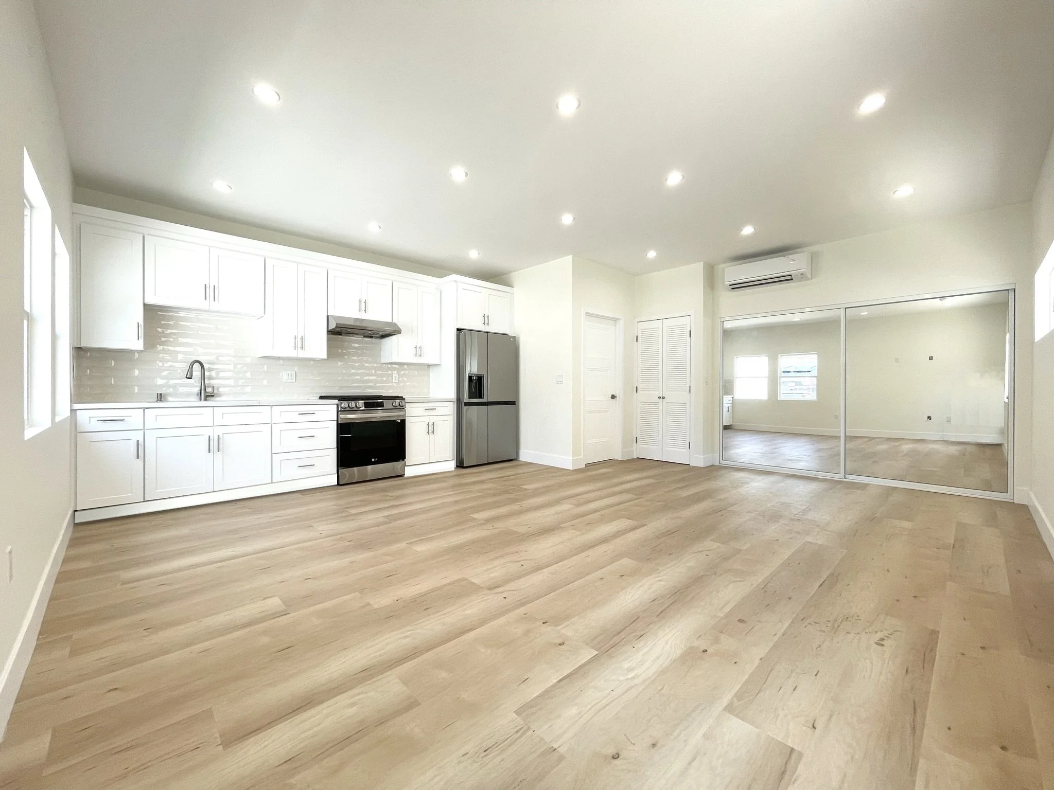 Open-concept kitchen and living area with white cabinets, stainless steel refrigerator, oven, and wood flooring, illuminated by recessed lighting.
