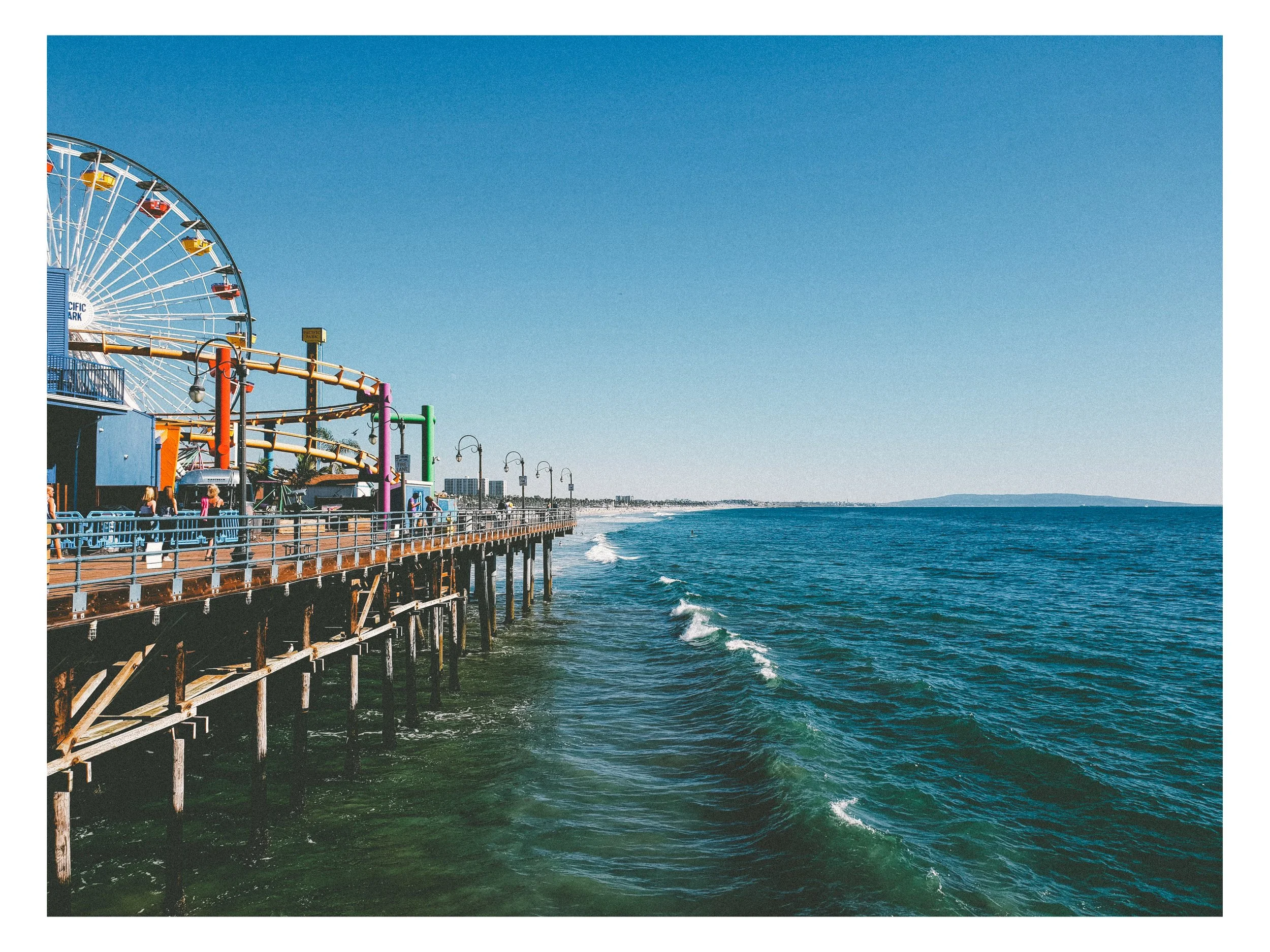 Pier extending into the ocean with a large Ferris wheel and colorful amusement park rides in the background, clear blue sky, and distant shoreline.