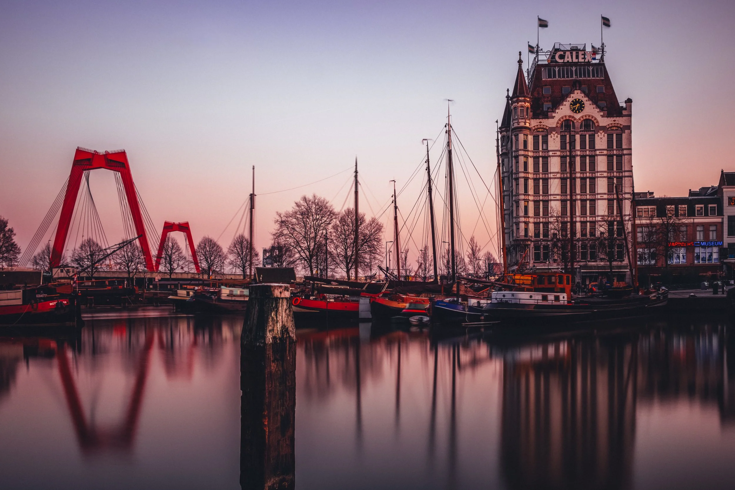 A harbor with several sailboats docked near a historic building at sunset. The water is calm and reflects the boats and the building. There are leafless trees and a large red bridge structure in the background.