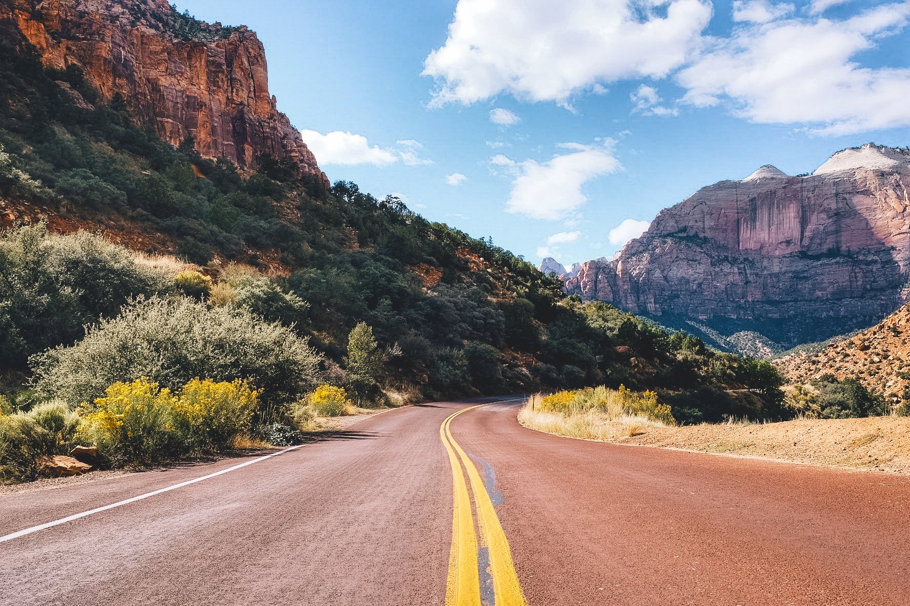 A winding road through a desert landscape with tall reddish rock formations and sparse vegetation under a partly cloudy sky.