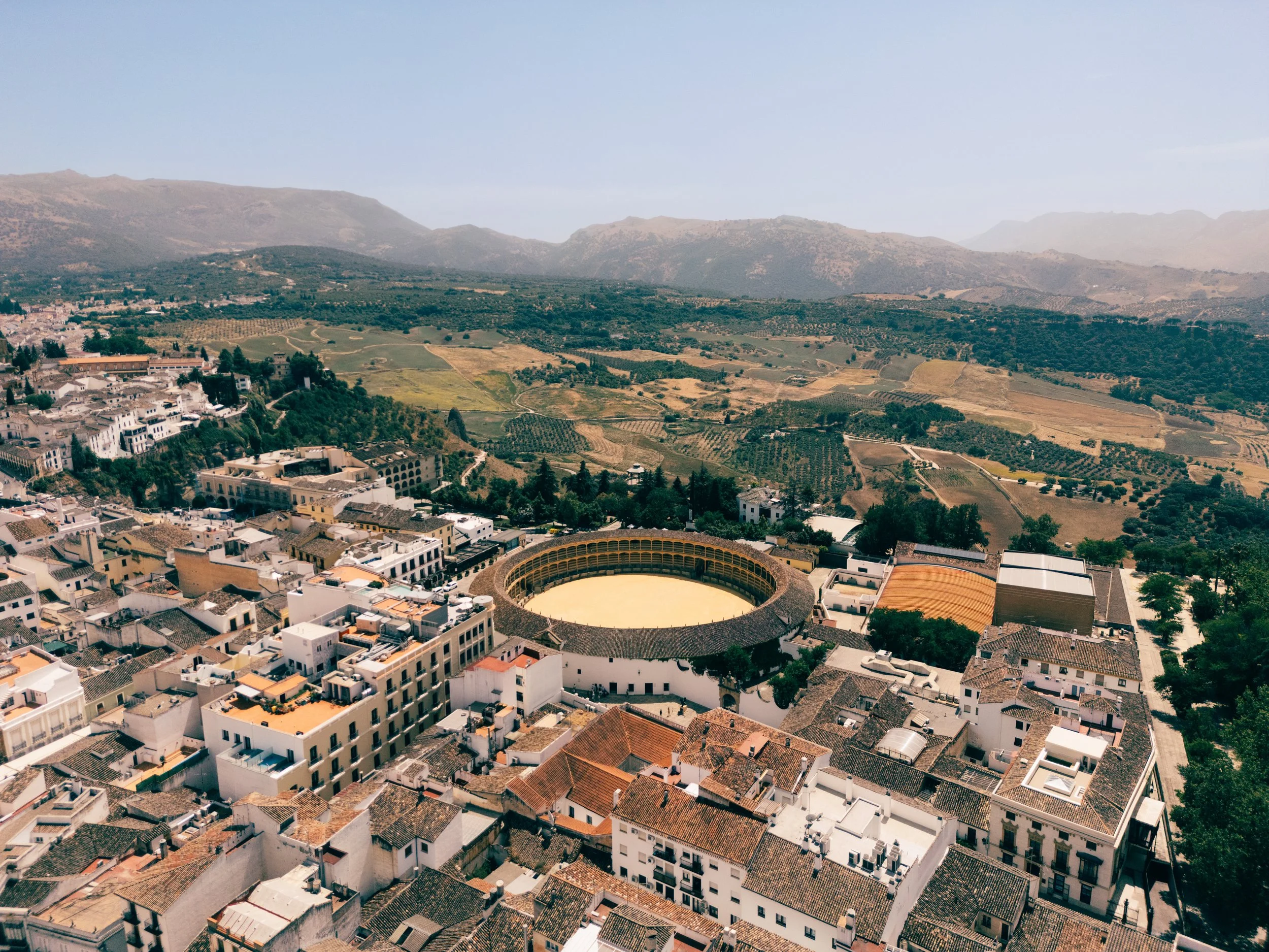 Aerial view of a city with a circular bullring and historic buildings, surrounded by green hills and mountains in the distance. Rhonda Spain, Spain landscape drone photography, Bull fighthing arena
