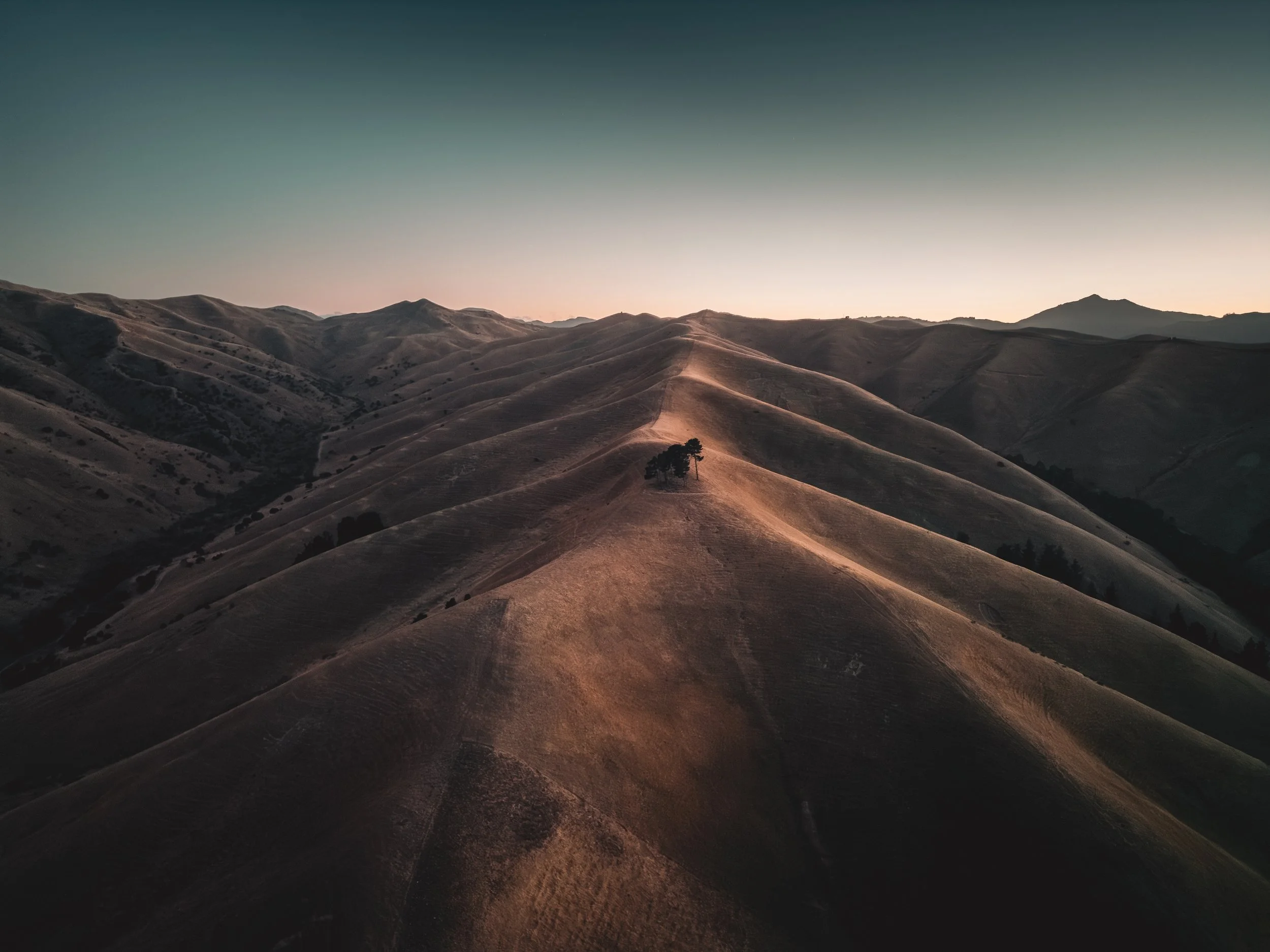A landscape of rolling hills at sunset with a few trees on a ridge, stretching into the distance with mountains on the horizon.