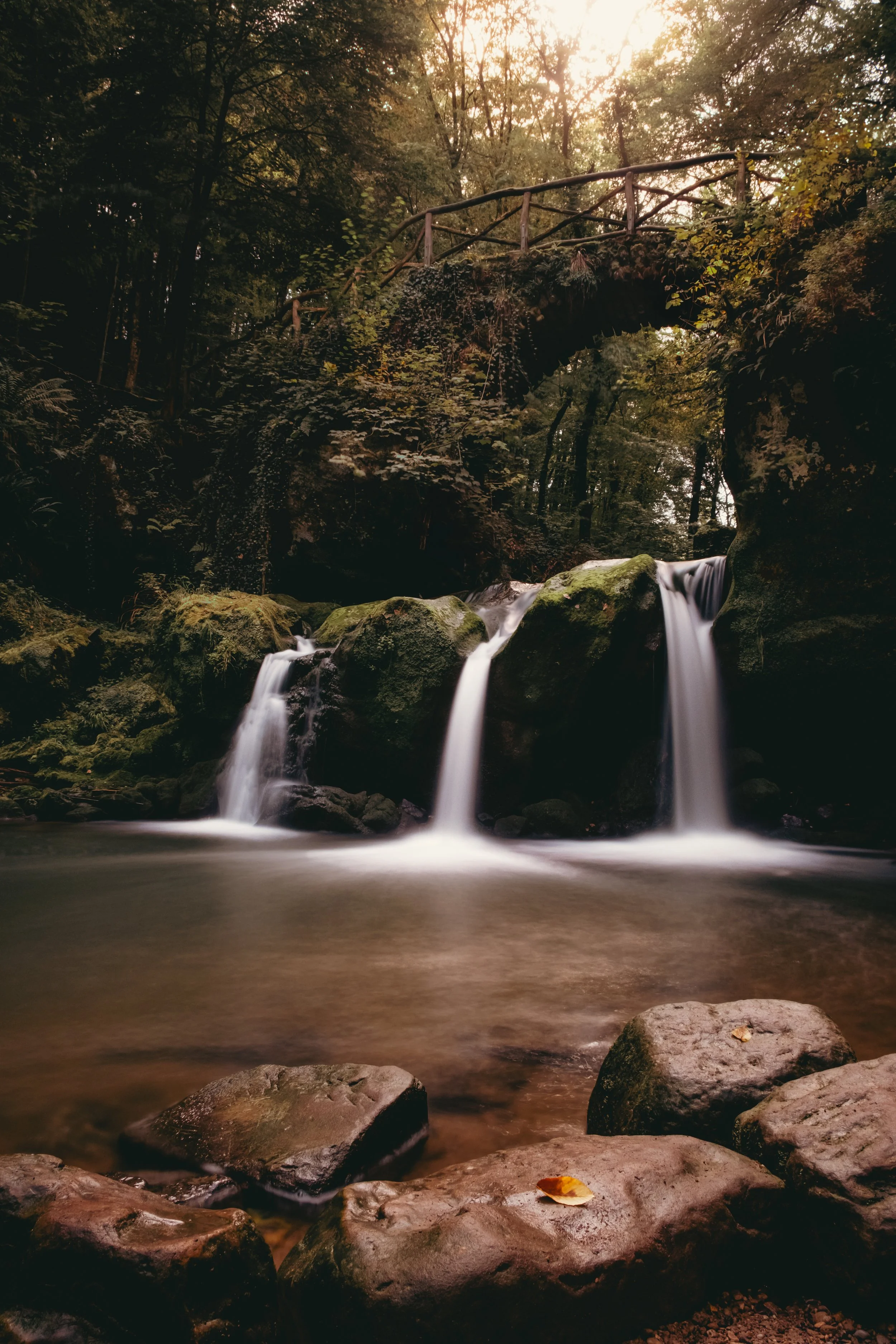 A serene forest scene with a small waterfall flowing over moss-covered rocks, a wooden bridge arching over the stream, and trees with sunlight filtering through the foliage.