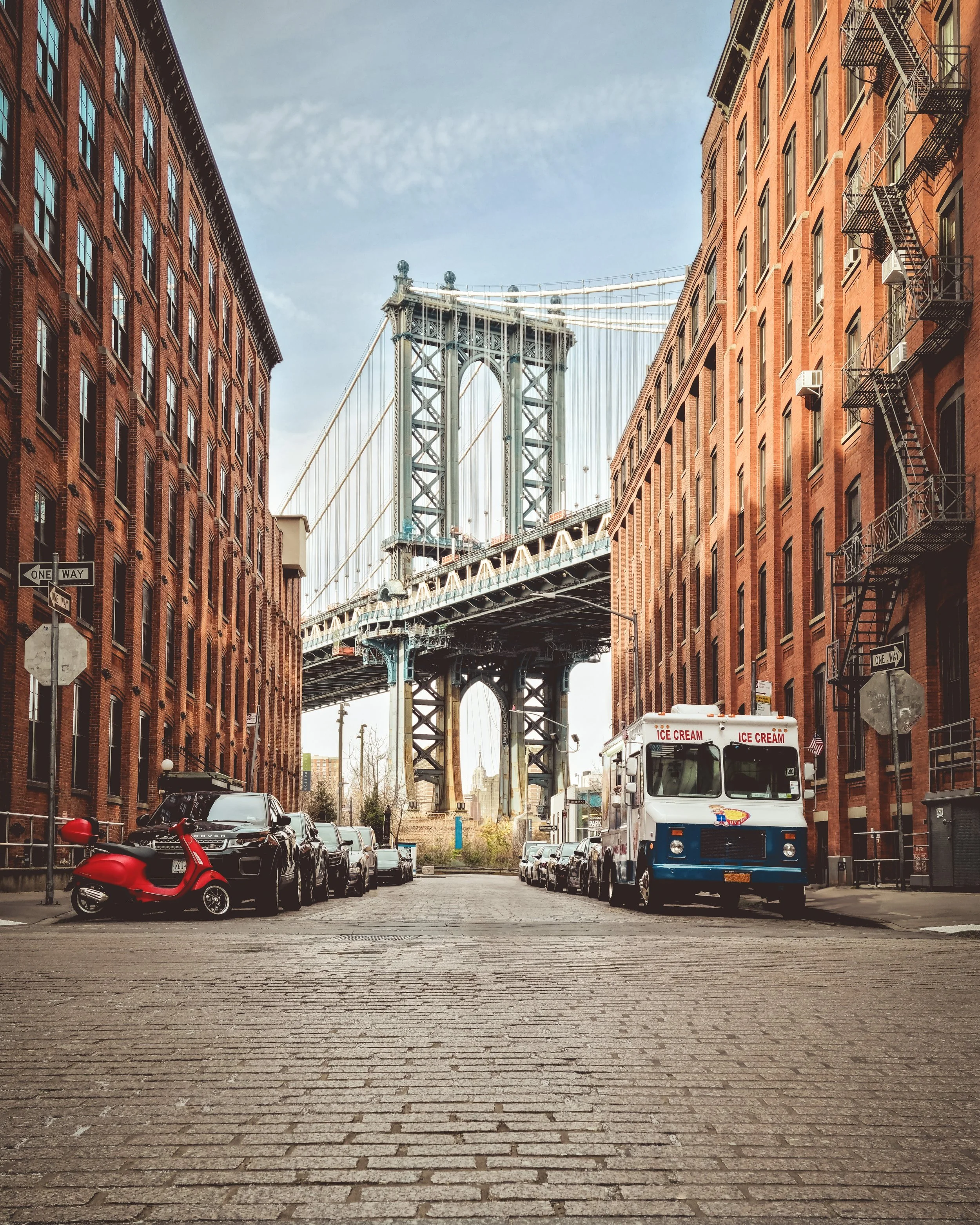 View of Manhattan Bridge through a cobblestone street framed by red brick buildings, with parked cars, an ice cream truck, a red scooter, and a few signs.