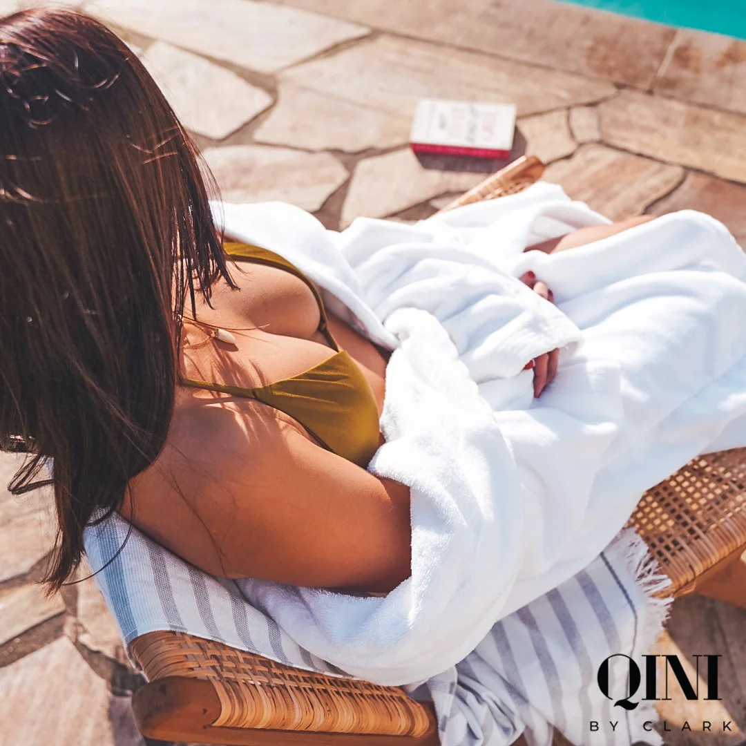 A woman in a green swimsuit lounging near a pool, partially covered with a white towel, with a white and gray striped towel on a wicker chair.