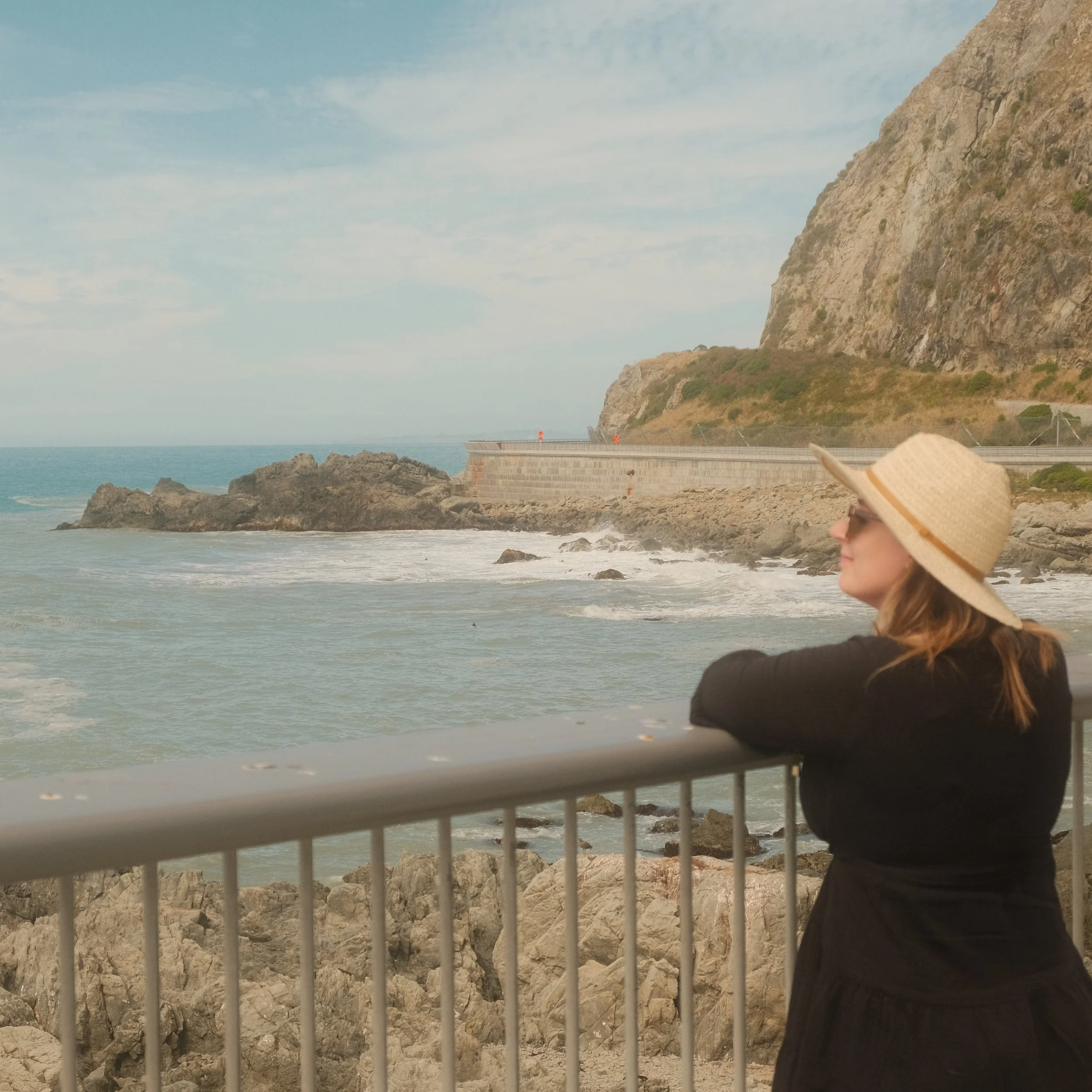 A woman wearing a large straw hat and sunglasses leaning on a metal railing while looking at the ocean and rocky coastline with cliffs in the background.