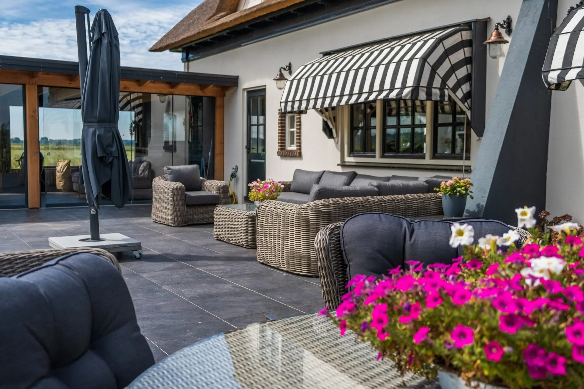 Outdoor patio with wicker and cushioned seating, potted flowers, an umbrella, a glass table, and a screened-in area in the background.